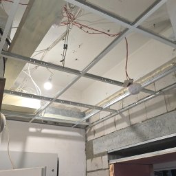 USŁUGI BUDOWLANO SPRZĄTAJĄCE R&B - Metalowa konstrukcja sufitu podwieszanego z widocznymi przewodami elektrycznymi i fragmentem ściany z betonu komórkowego. Etap prac wykończeniowych.