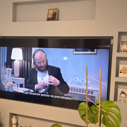 USŁUGI BUDOWLANO SPRZĄTAJĄCE R&B - Ściana z szarej płyty KG z wnękami, w których umieszczono zdjęcia w ramkach. Na ścianie wisi telewizor. Dekoracja ściany z płyt kartonowo-gipsowych.