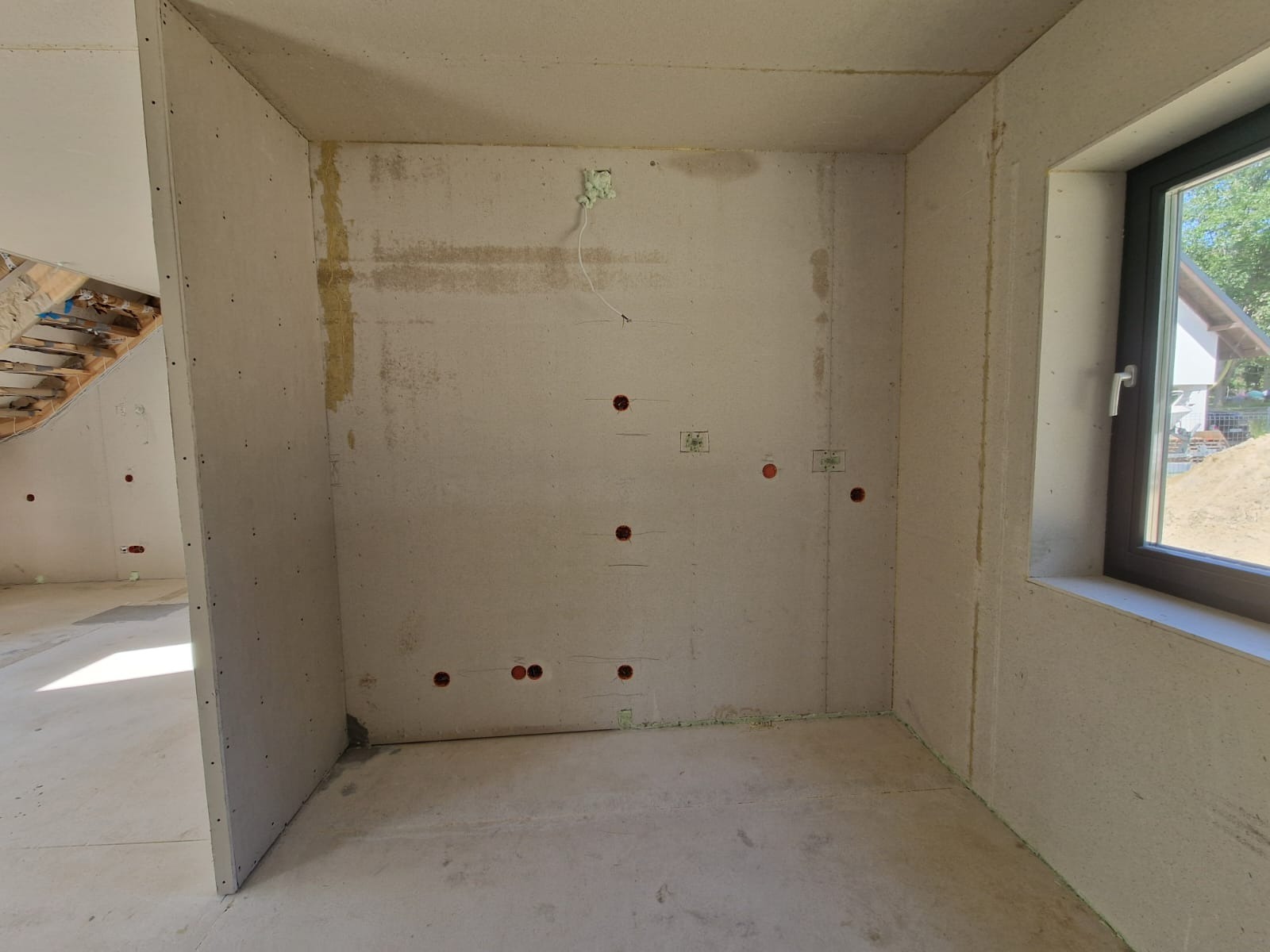 Wnętrze pokoju w trakcie wykańczania, ściany obłożone płytami gipsowo-kartonowymi, widoczne otwory na instalacje elektryczne i fragment okna z widokiem na zewnątrz.