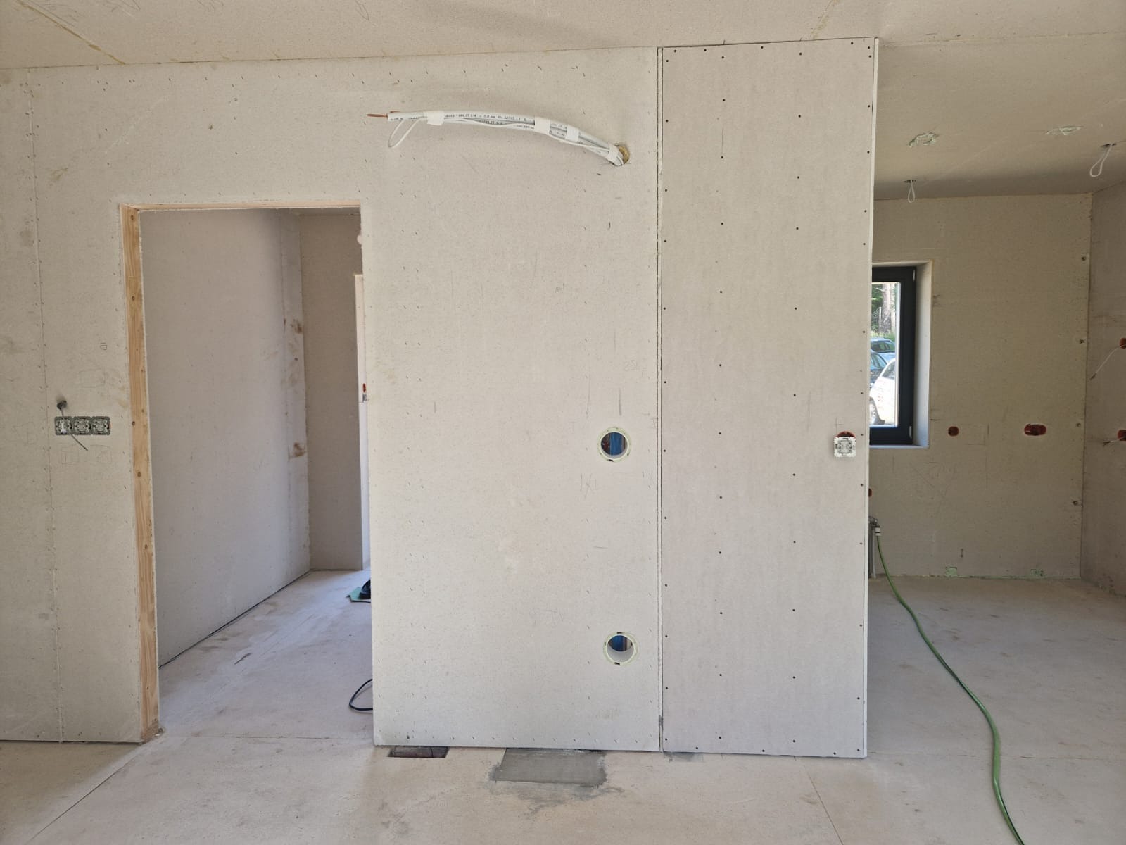 Wnętrze w trakcie prac wykończeniowych: ściany obłożone płytami gipsowo-kartonowymi z widocznymi otworami na instalacje, surowy betonowy podkład, wystające przewody elektryczne i instalacyjne.