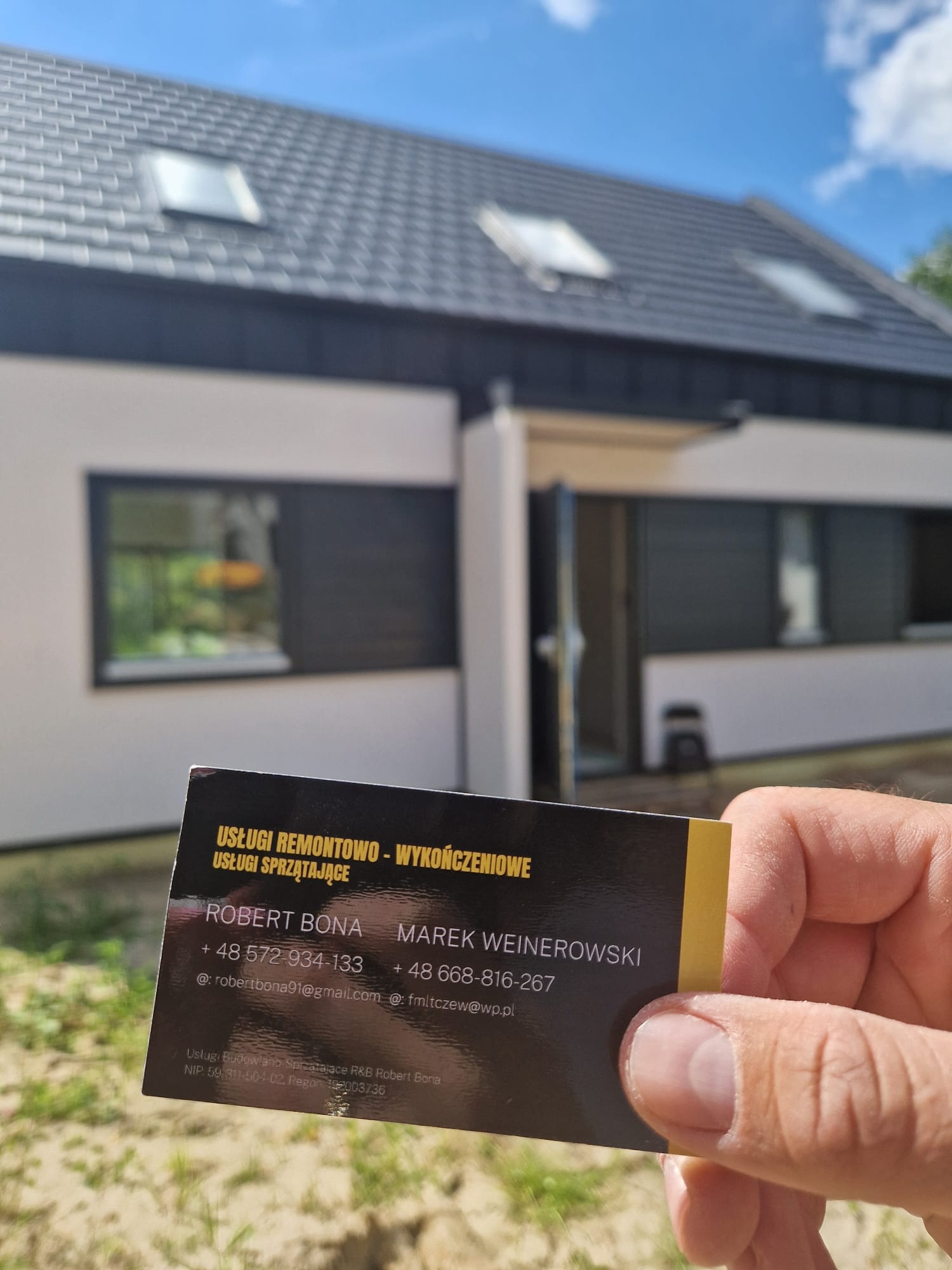 Wizytówka firmy remontowo-wykończeniowej na tle nowo wybudowanego domu z szarym dachem i białymi ścianami, z oknami z czarnymi ramami.