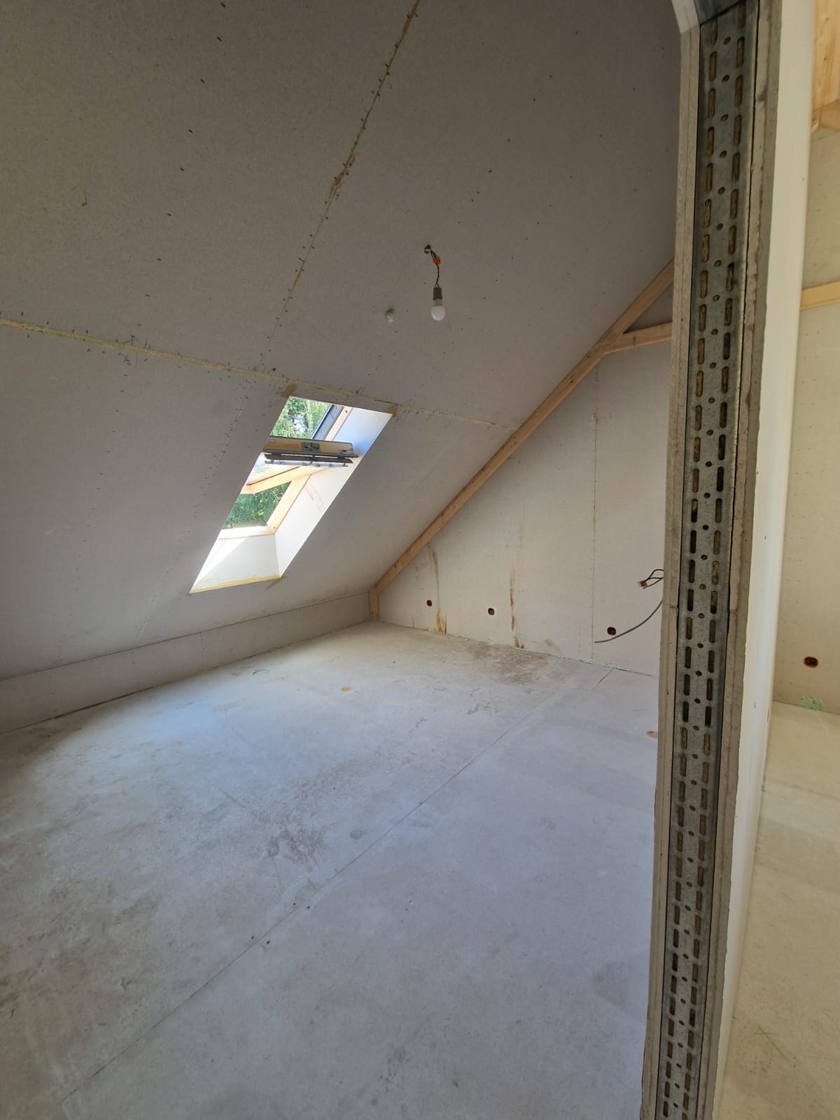 Poddasze w trakcie wykańczania, widoczne płyty na skosach dachu, okno dachowe z widokiem na zieleń, surowa podłoga i metalowa konstrukcja ściany działowej.