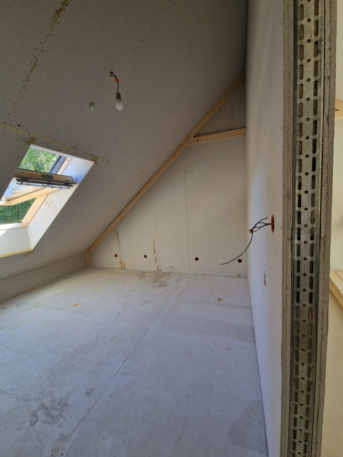 Poddasze w trakcie wykańczania: widoczne surowe ściany z płyt, drewniana konstrukcja dachu, okno dachowe i wystające kable elektryczne z puszki w ścianie.
