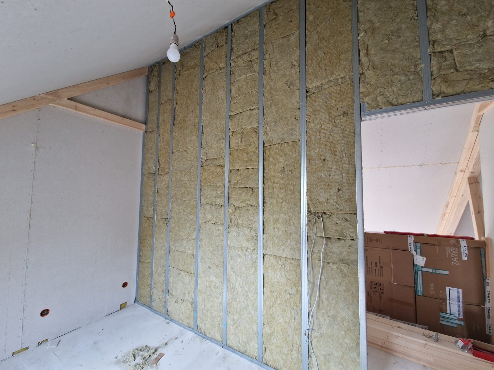 Montaż izolacji z wełny mineralnej w metalowej konstrukcji szkieletowej, widoczne przewody elektryczne, częściowo zamontowana płyta na ścianie, surowy stan wykończenia wnętrza.