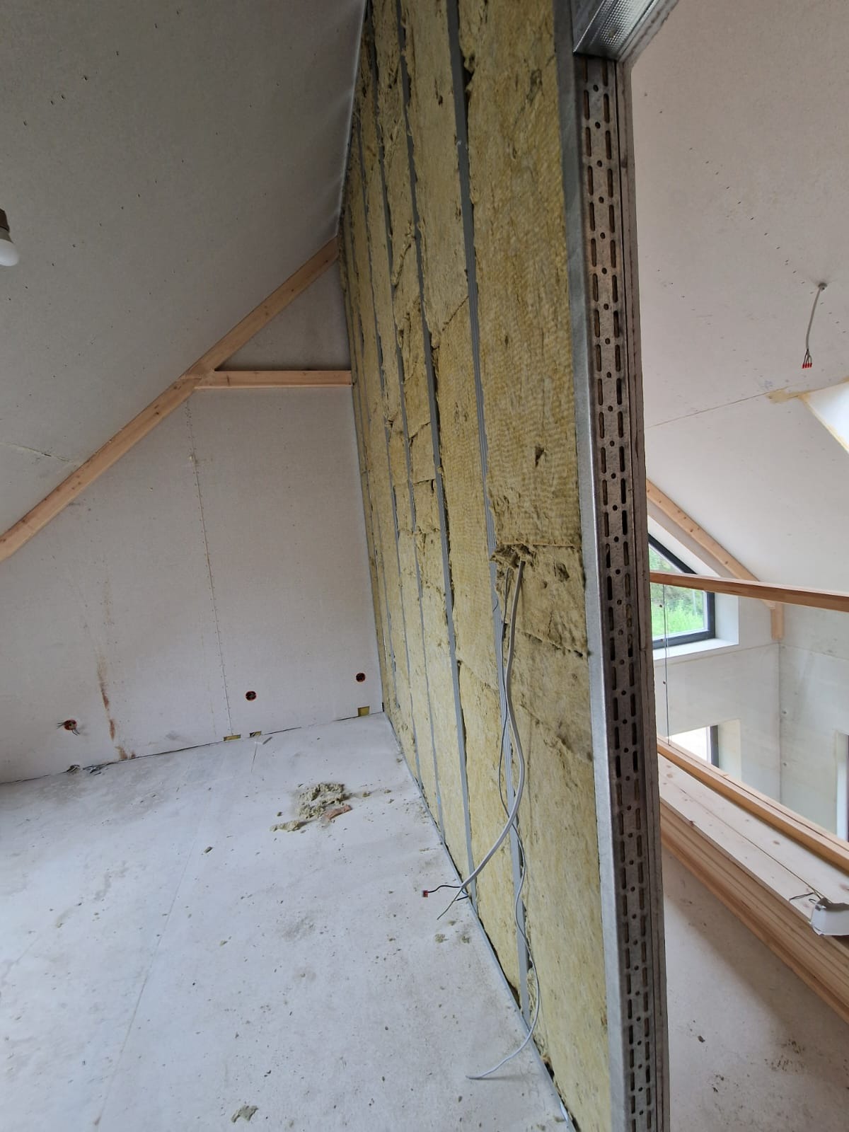 Montaż izolacji z wełny mineralnej w metalowym stelażu na poddaszu, widoczne przewody elektryczne i fragment konstrukcji dachu