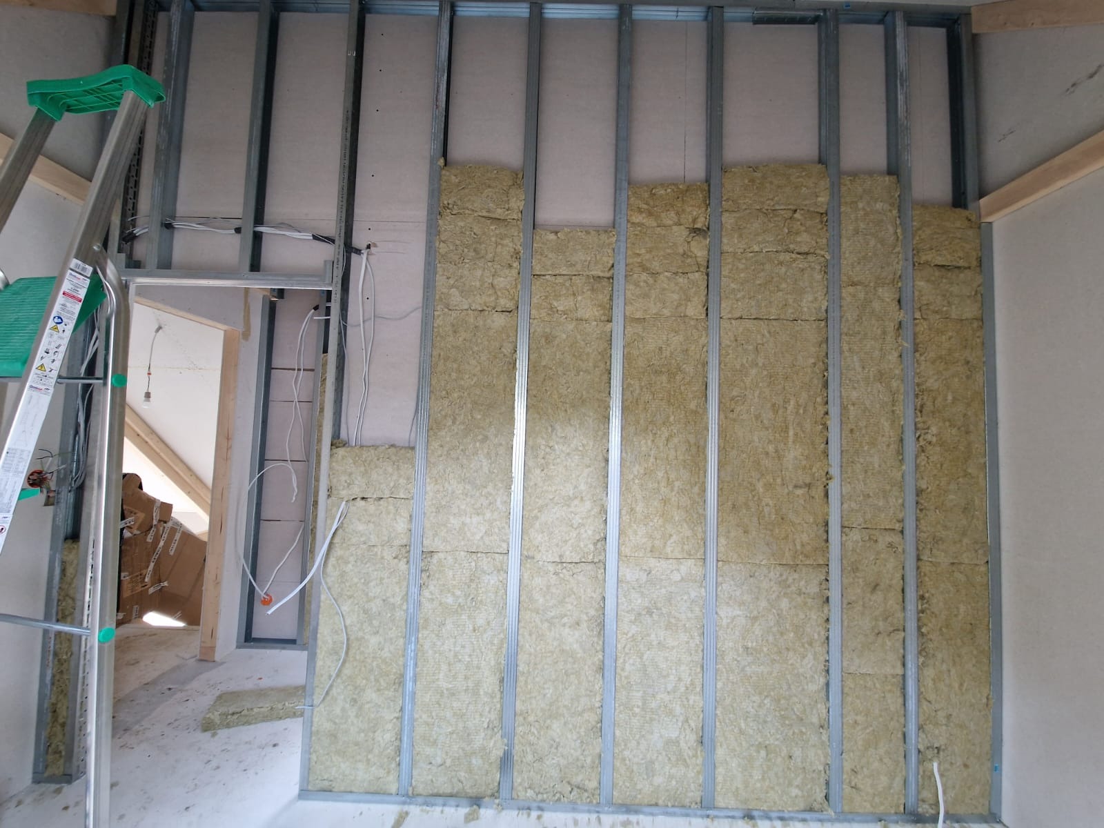 Wnętrze w trakcie prac wykończeniowych: metalowy szkielet ściany z częściowo wypełnioną wełną mineralną, widoczna drabina i przewody elektryczne.
