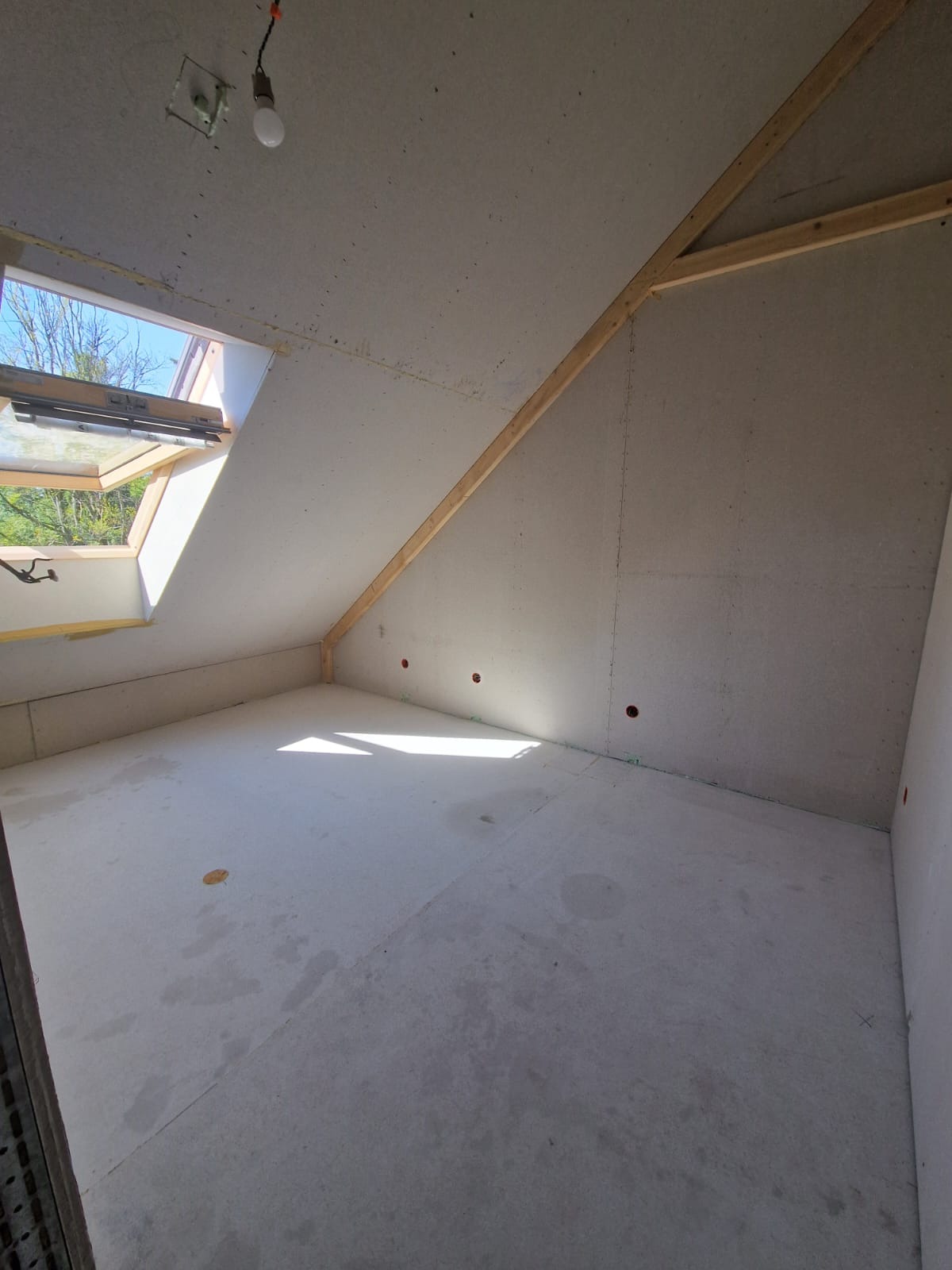 Poddasze z oknem dachowym w trakcie wykańczania, ściany obłożone płytami, widoczne drewniane belki konstrukcyjne i instalacja elektryczna.