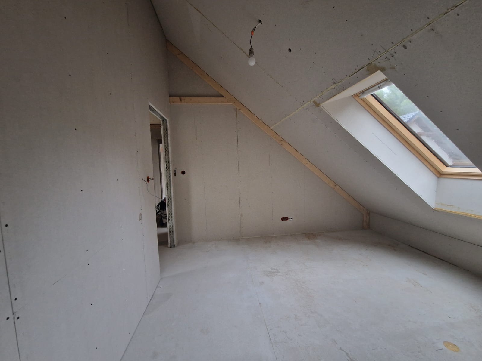 Pokój na poddaszu w trakcie remontu, ściany i skosy wyłożone płytami karton gips, widoczne drewniane elementy konstrukcyjne, okno dachowe i wisząca żarówka.