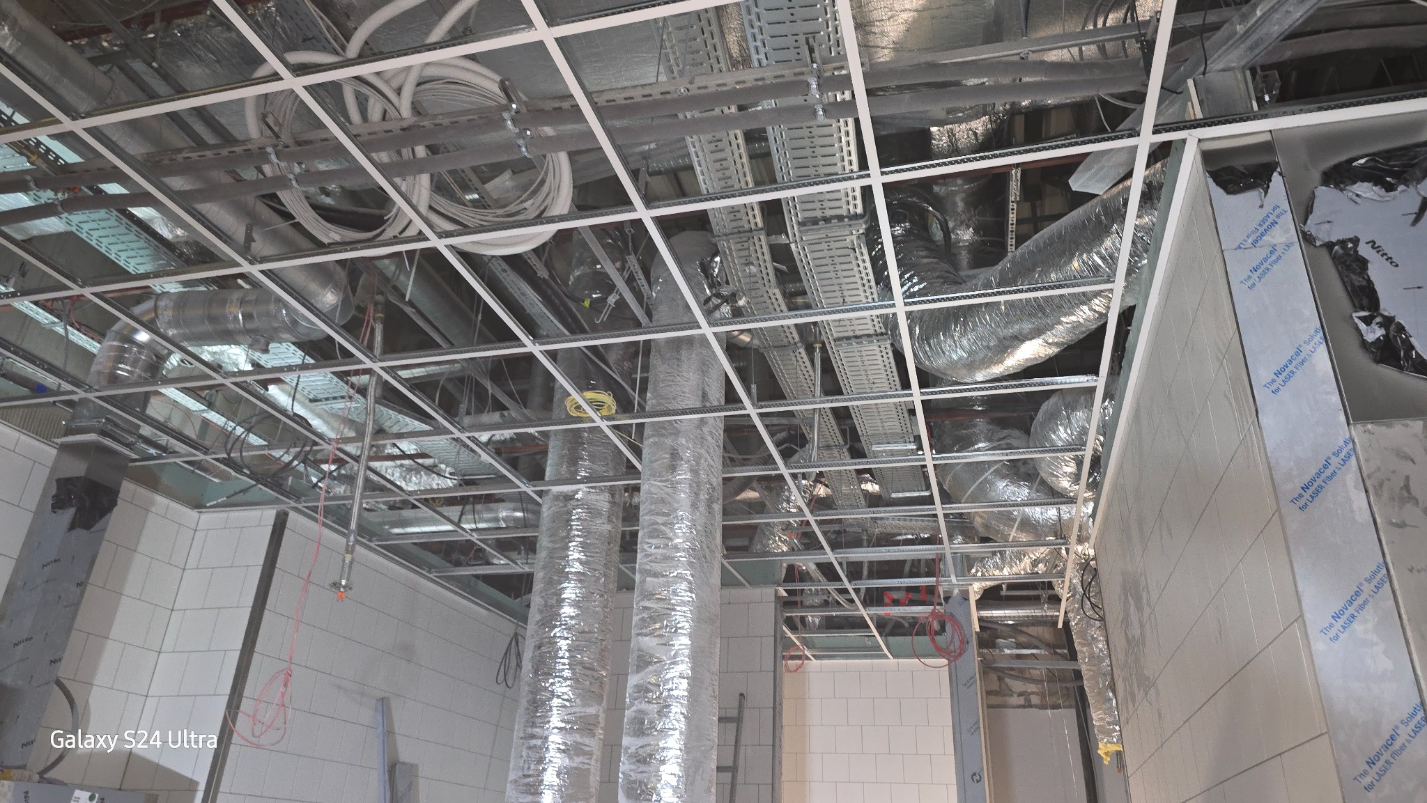 Widok na sufit podwieszany z widocznymi elementami instalacji wentylacyjnej, przewodami elektrycznymi i elementami konstrukcyjnymi, w trakcie montażu w pomieszczeniu z białymi płytkami ściennymi.