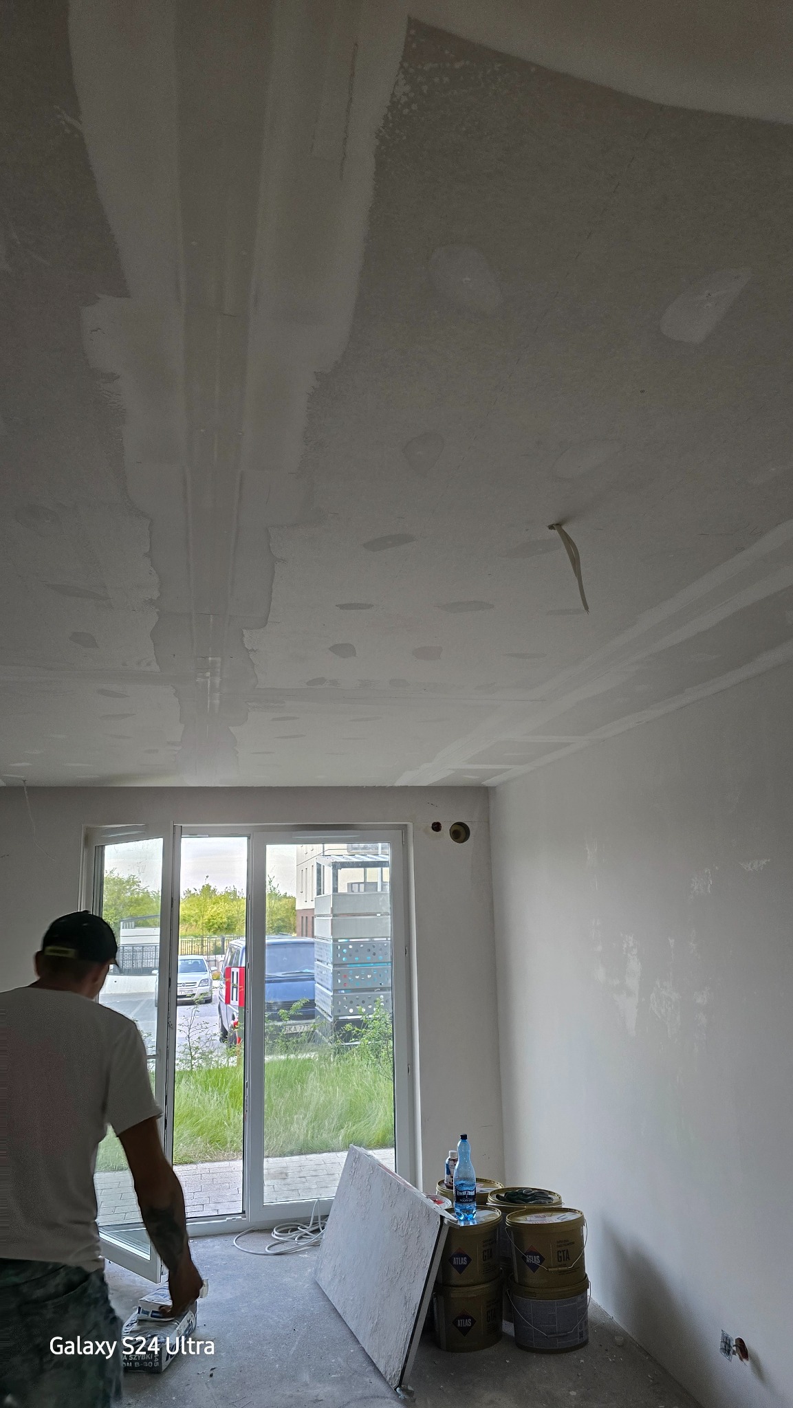 Wnętrze w trakcie wykańczania: widok na sufit ze śladami po łataniu i szpachlowaniu, ściany przygotowane pod malowanie, widoczne wiadra z materiałami budowlanymi, pracownik w tle w pobliżu okna.