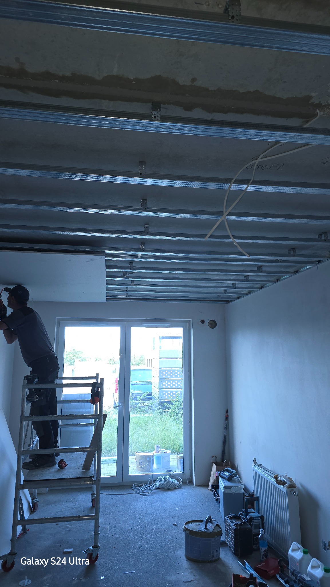 Pracownik na rusztowaniu montuje płytę na metalowym stelażu sufitowym w pomieszczeniu z oknem balkonowym, widoczne narzędzia i materiały budowlane.