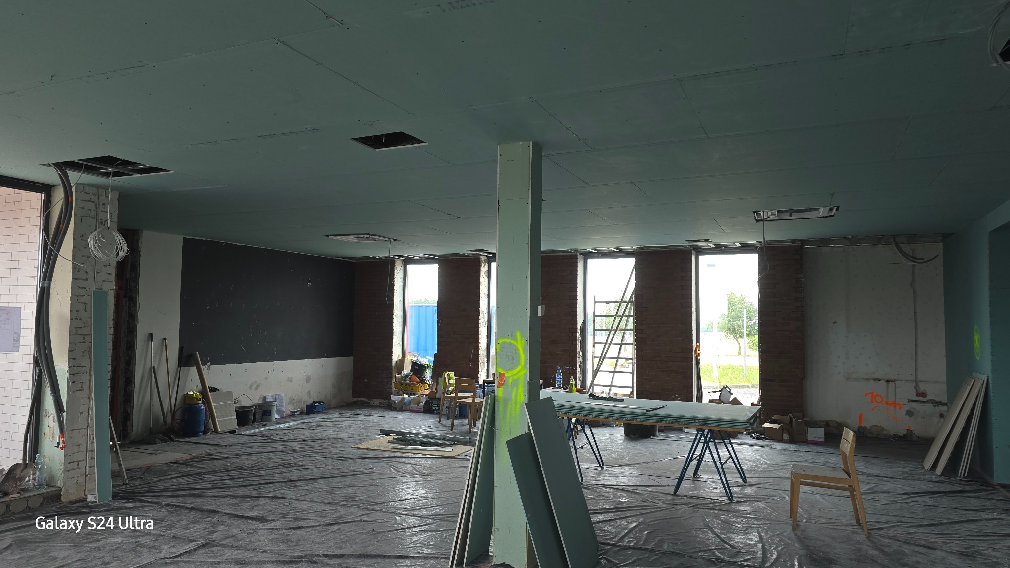 Wnętrze w trakcie remontu: surowy strop pokryty płytami, widoczne instalacje, narzędzia, folia ochronna na podłodze i fragmenty ceglanej ściany w tle.