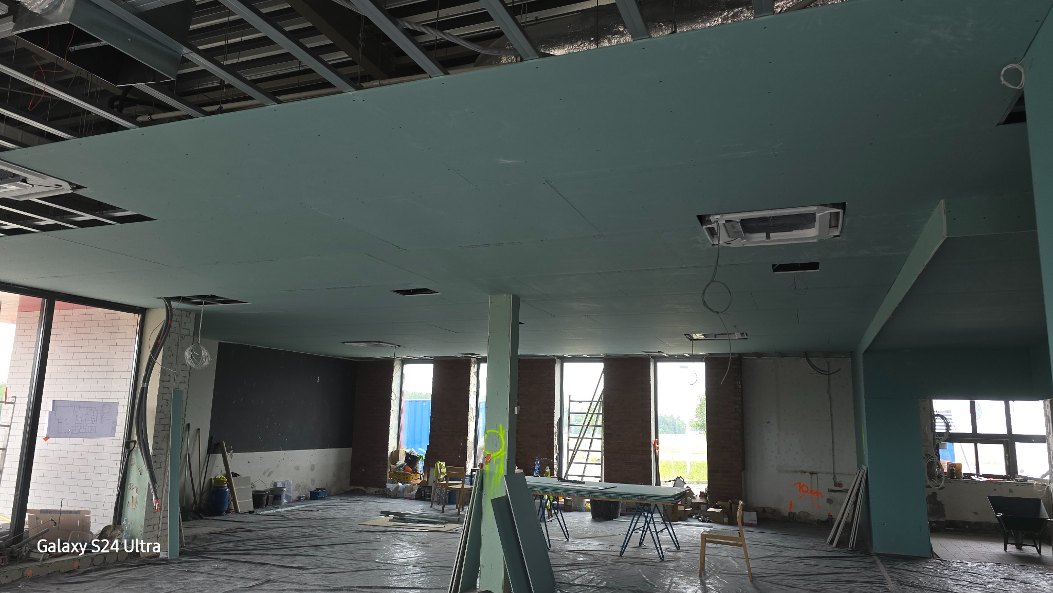 Montaż sufitu podwieszanego z płyt karton-gips w dużym pomieszczeniu biurowym, widoczne profile konstrukcyjne, otwory na oświetlenie i instalacje wentylacyjne, podłoga zabezpieczona folią.