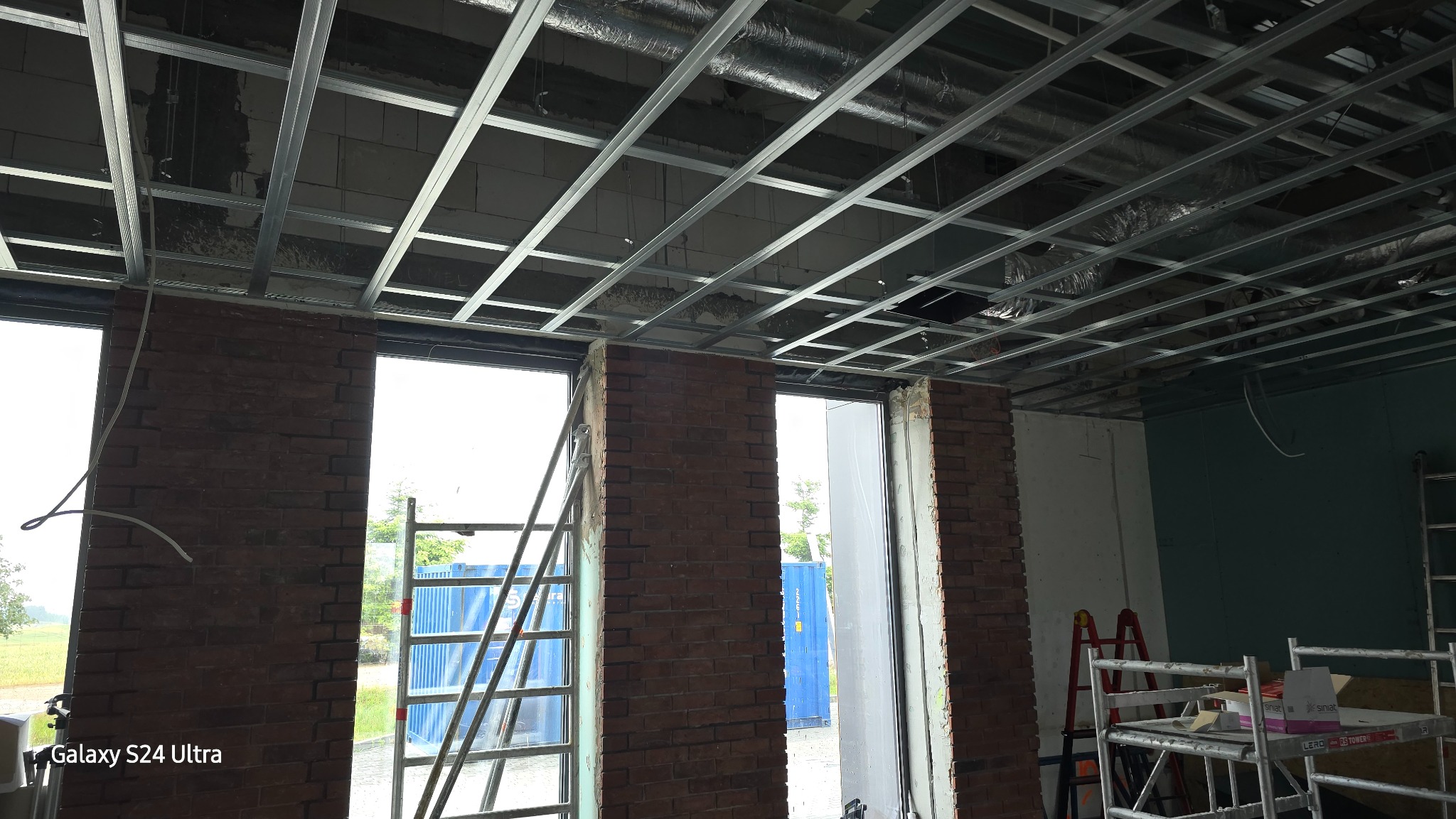 Wnętrze w trakcie remontu: widoczne rusztowanie metalowe sufitu podwieszanego, fragmenty ścian z cegły klinkierowej, drabina oparta o ścianę, okna z widokiem na zewnątrz i elementy wyposażenia...