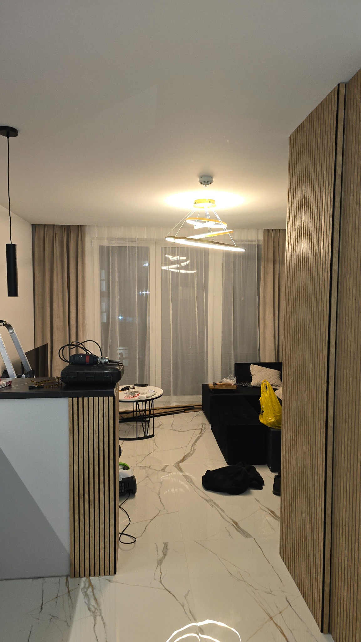 Nowoczesne wnętrze z marmurową podłogą i panelami ściennymi z drewna. Widoczny fragment sofy, stolik kawowy i designerska lampa sufitowa. W tle okno z zasłonami.