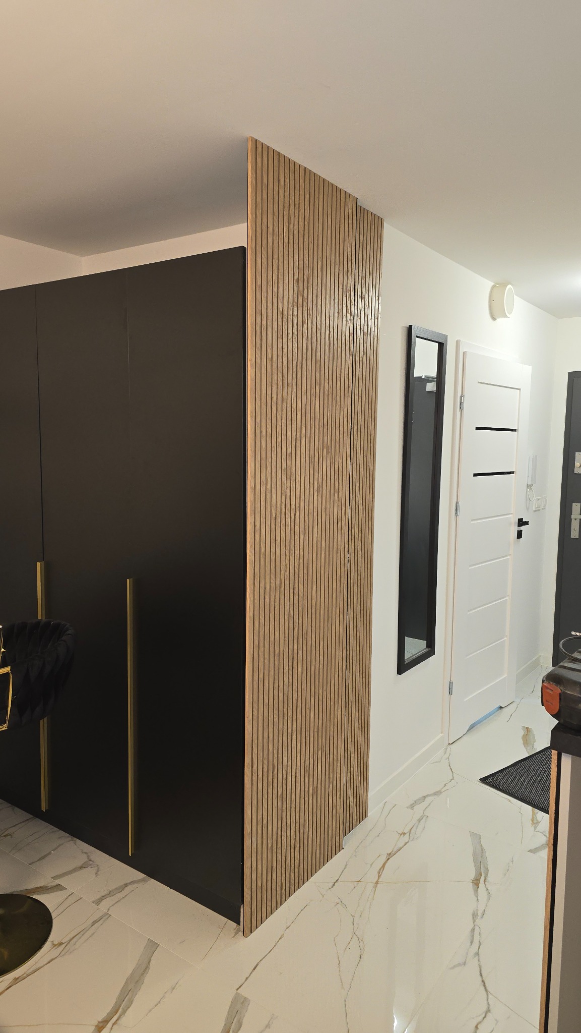 Nowoczesne wnętrze z drewnianą ścianką działową z lameli, czarną szafą z złotymi uchwytami i marmurową podłogą. Minimalistyczny design z lustrem i białymi drzwiami w tle.