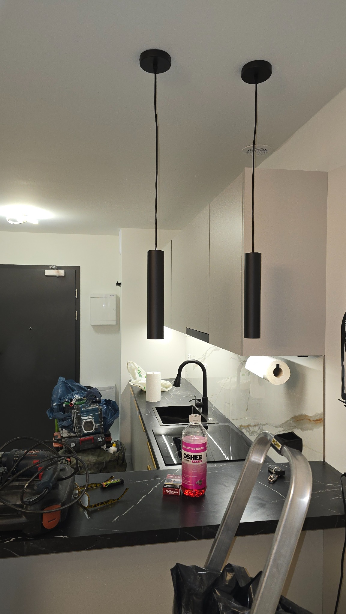 Nowoczesna kuchnia w trakcie montażu: czarne lampy cylindryczne nad blatem z marmuru, widoczne narzędzia i drabina, sugerujące etap wykończeniowy.
