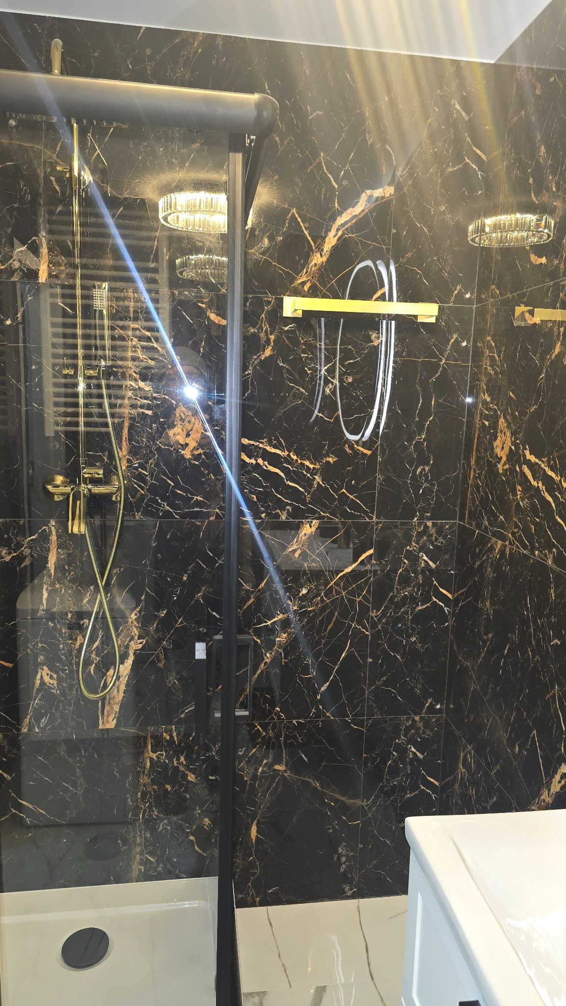 Elegancka kabina prysznicowa z czarnymi płytkami imitującymi marmur ze złotymi żyłami, złota armatura i oświetlenie. Nowoczesny design łazienki.
