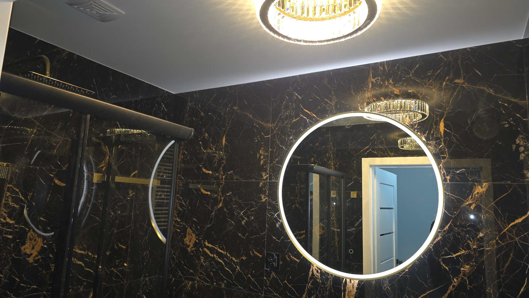 Elegancka łazienka z czarnymi płytkami imitującymi marmur, okrągłe lustro z podświetleniem LED i kabina prysznicowa z czarnymi profilami. Lampa sufitowa kryształowa.