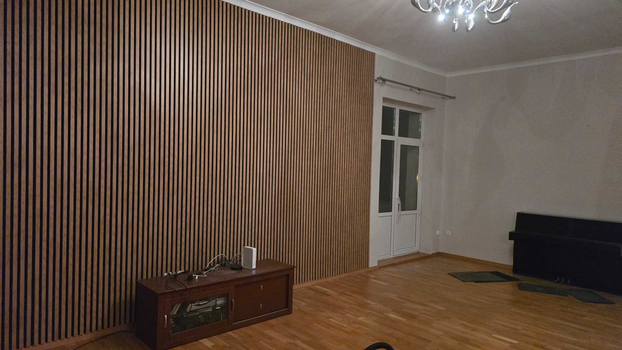 Ściana wykończona panelami lamelowymi w odcieniach brązu, tworzącymi pionowy wzór. Widoczny fragment pokoju z parkietem, szafką RTV i drzwiami balkonowymi.