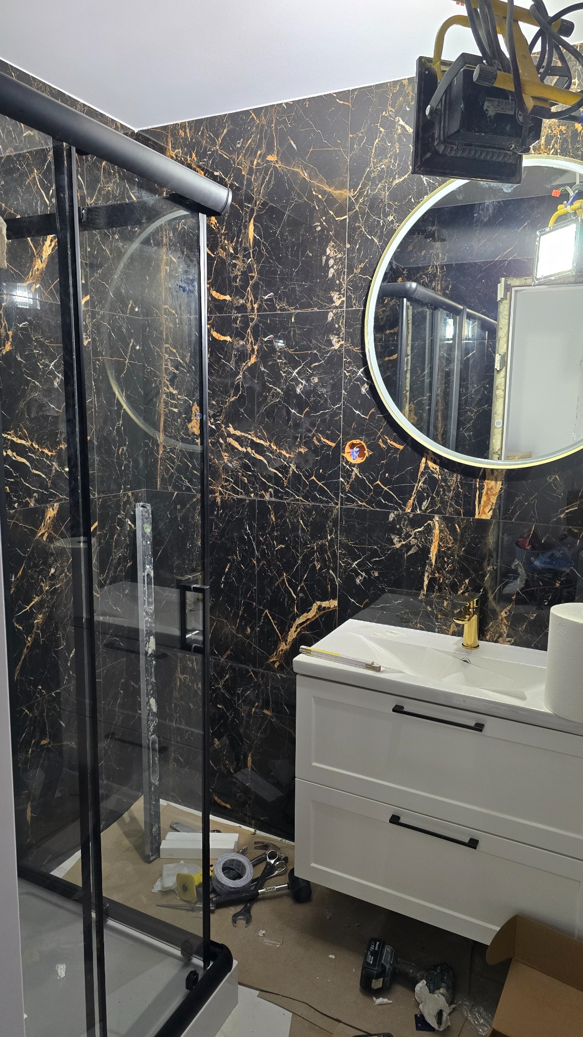 Nowoczesna łazienka z czarnymi płytkami imitującymi marmur, kabiną prysznicową z czarnymi ramami i białą szafką pod umywalką. Widoczne narzędzia sugerują prace wykończeniowe.