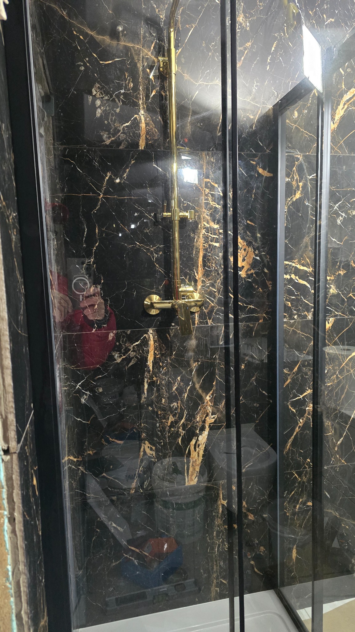Kabina prysznicowa z czarnymi płytkami imitującymi marmur ze złotymi żyłami i złotą armaturą. Widoczne odbicie osoby robiącej zdjęcie.