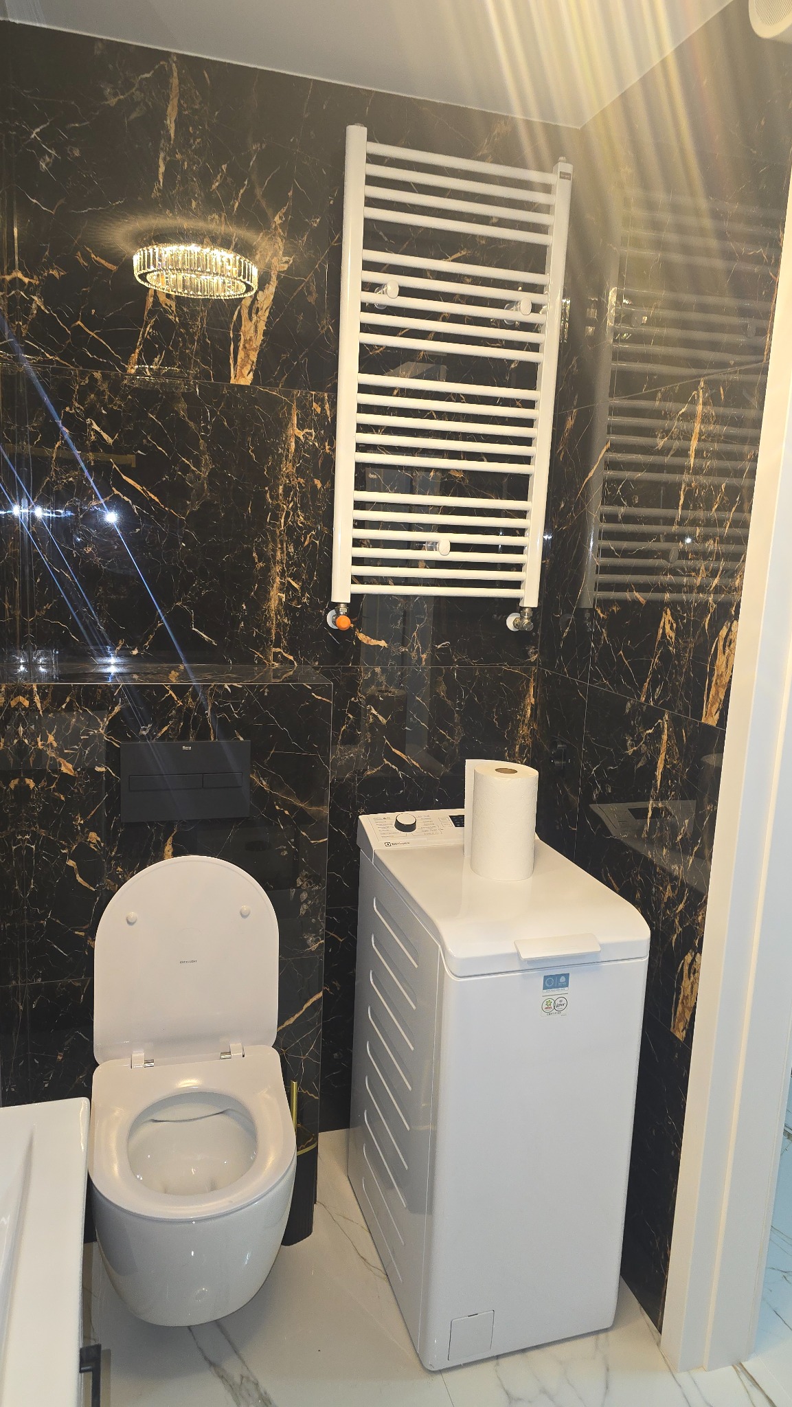 Elegancka łazienka z czarnymi płytkami imitującymi marmur, białą toaletą, pralką i grzejnikiem drabinkowym. Kryształowy plafon dodaje luksusowego charakteru.