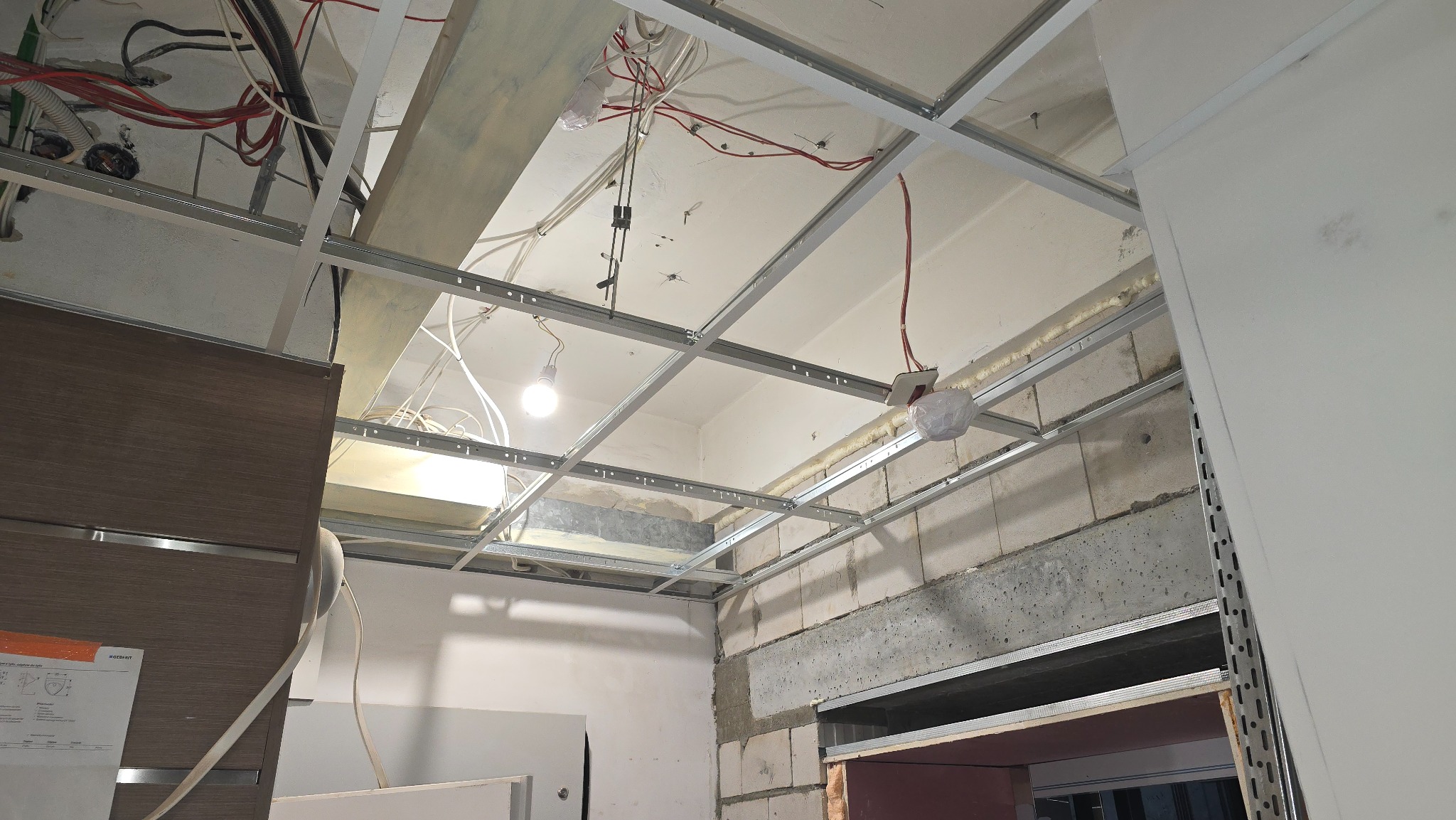 Metalowa konstrukcja sufitu podwieszanego z widocznymi przewodami elektrycznymi i fragmentem ściany z betonu komórkowego. Etap prac wykończeniowych.