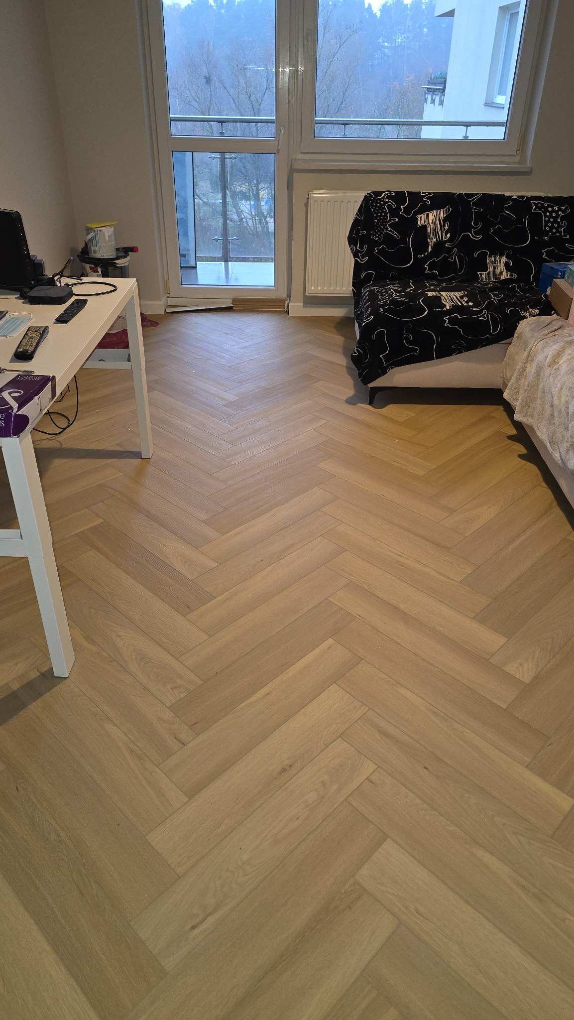 Jasna podłoga z paneli ułożonych w jodełkę francuską, widok na balkon i fragment pokoju z biurkiem i sofą. Naturalne światło podkreśla wzór podłogi.