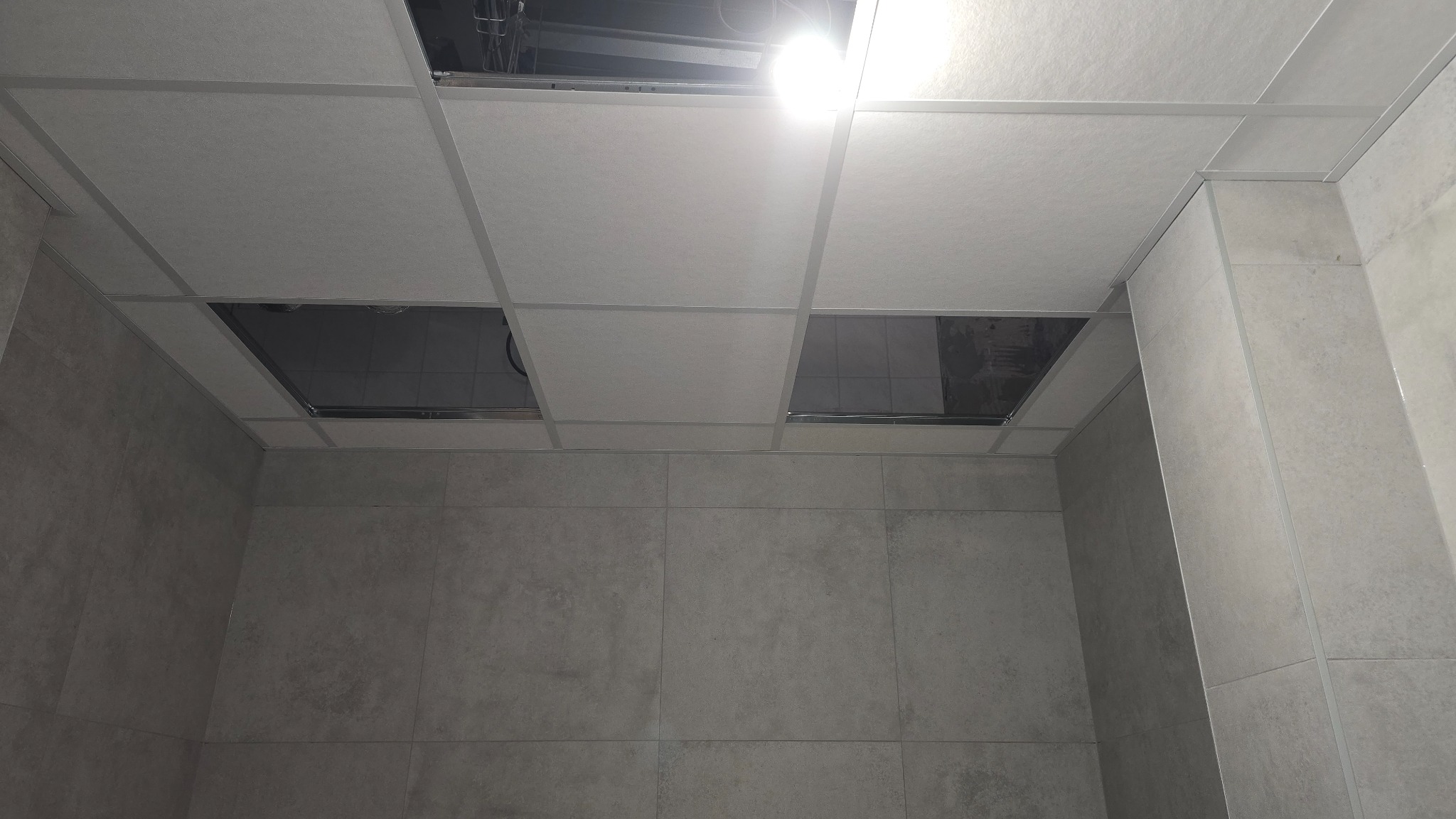 Kwadratowa, podwieszana sufitowa konstrukcja z widocznymi otworami i oświetleniem, połączona z wykończoną, szarą, ceramiczną ścianą w łazience.