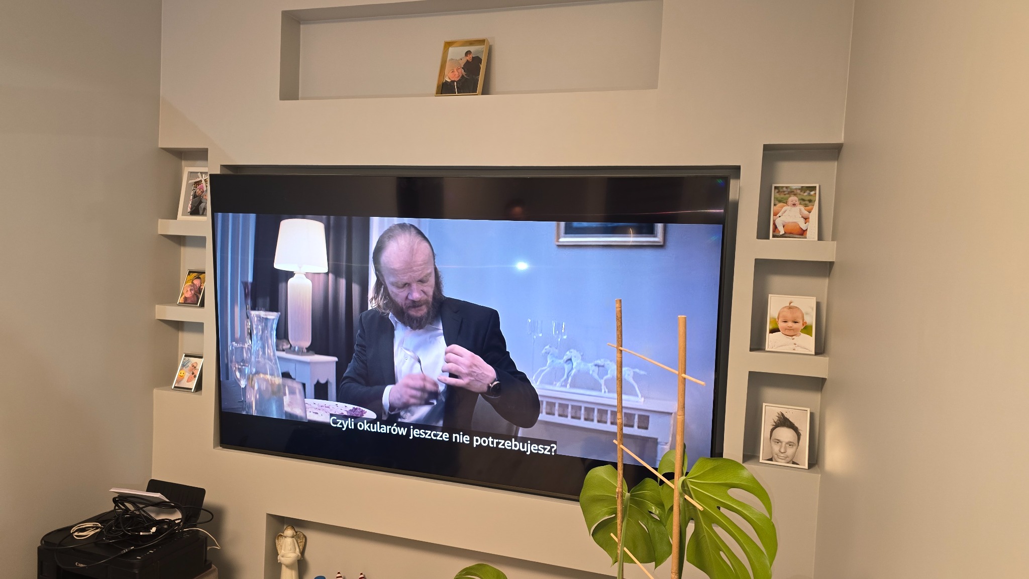 Ściana z szarej płyty KG z wnękami, w których umieszczono zdjęcia w ramkach. Na ścianie wisi telewizor. Dekoracja ściany z płyt kartonowo-gipsowych.