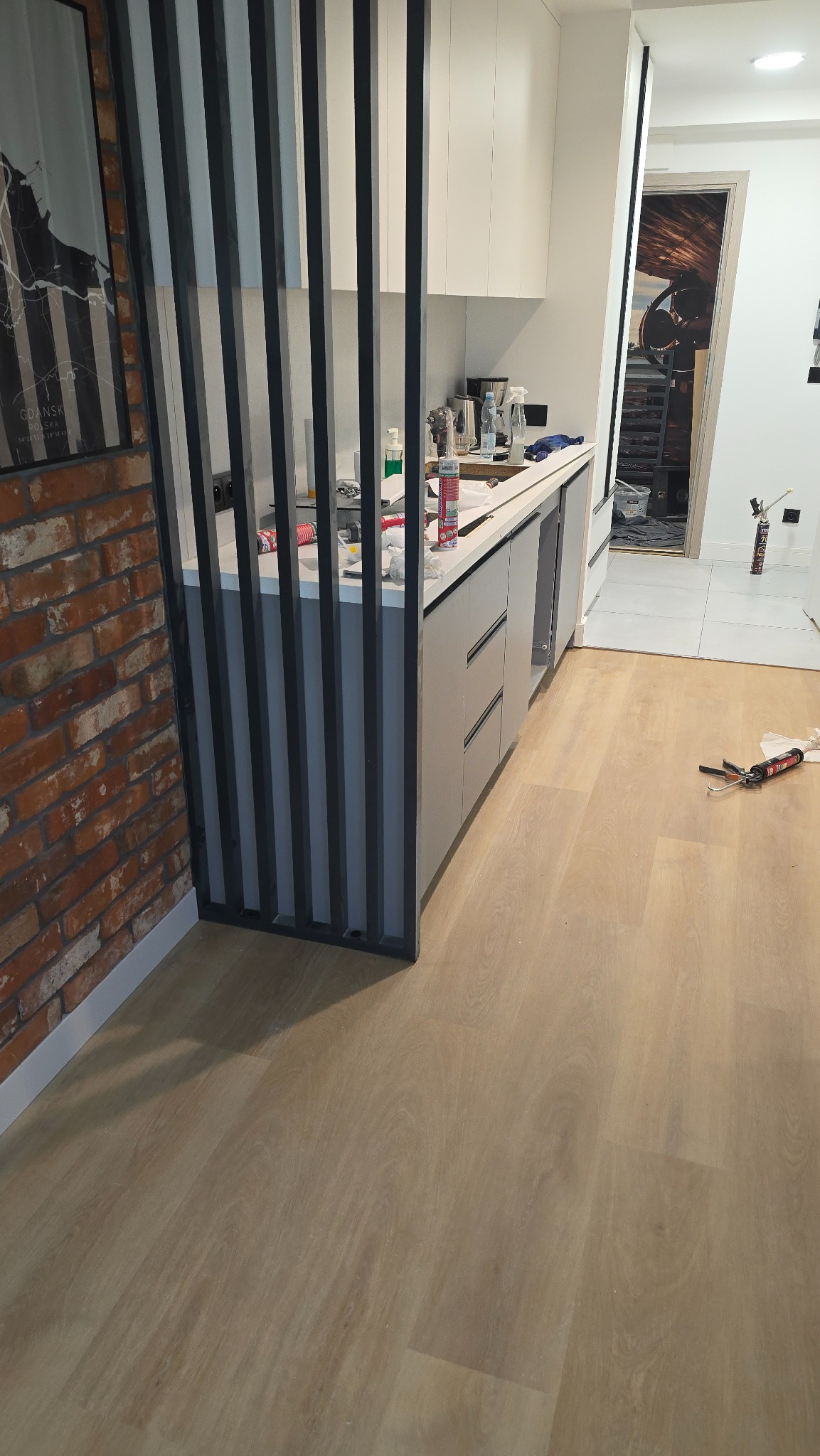 Nowoczesna kuchnia z drewnianą podłogą i ceglaną ścianą, oddzielona ażurową ścianką. Widoczne narzędzia i materiały budowlane sugerują prace wykończeniowe.