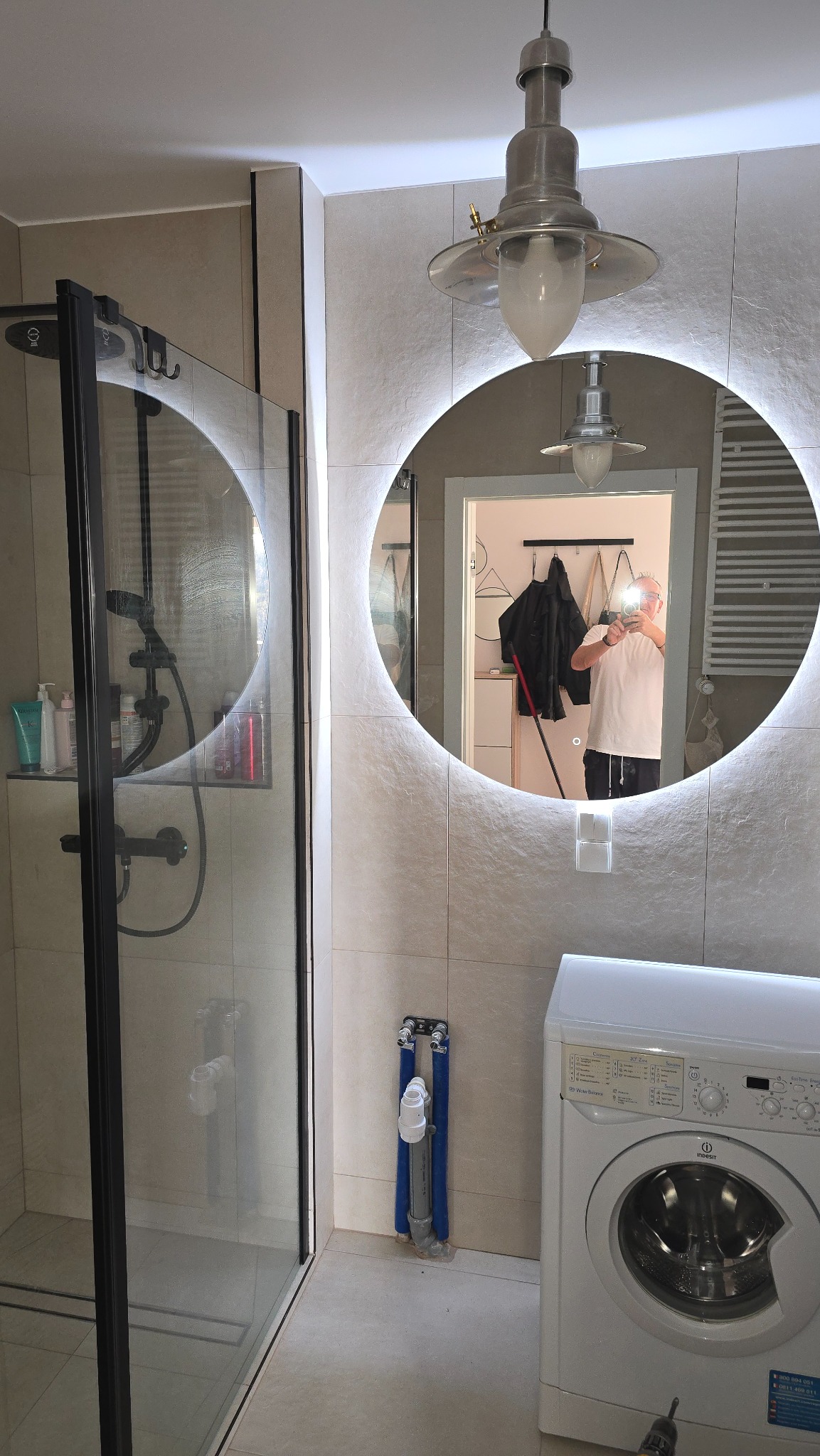 Nowoczesna łazienka z kabiną prysznicową, okrągłym lustrem z podświetleniem LED i pralką. Widoczny fragment instalacji hydraulicznej przy kabinie.