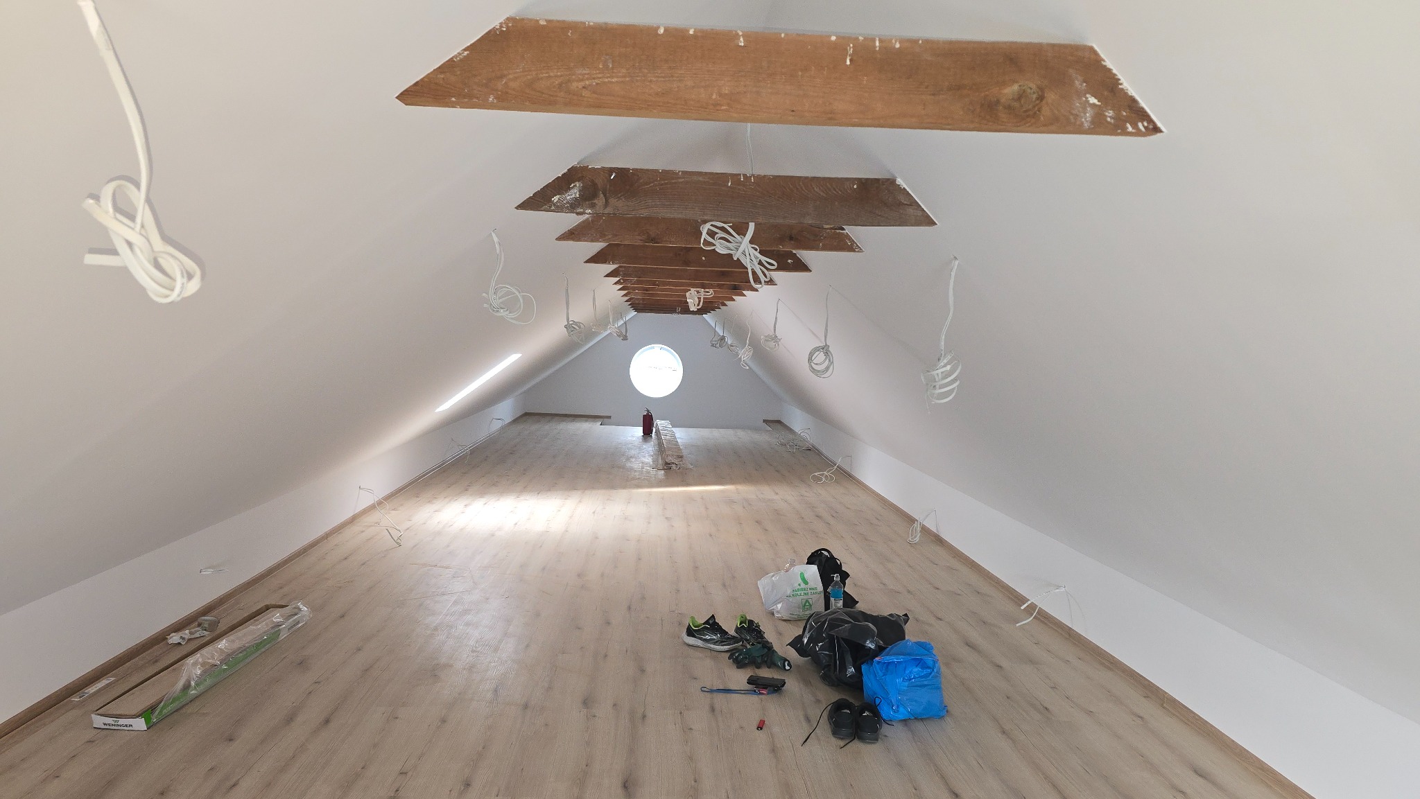 Poddasze z drewnianymi belkami i oknem. Na podłodze panele, narzędzia i kable zwisające z sufitu. Widok na ukończone układanie paneli podłogowych.