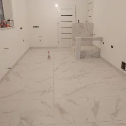 Jasna łazienka w trakcie remontu, z położoną marmurkową podłogą z płytek i fragmentem obudowy wanny lub brodzika z tych samych płytek, widoczne puszki instalacyjne na ścianach i butelka ze...