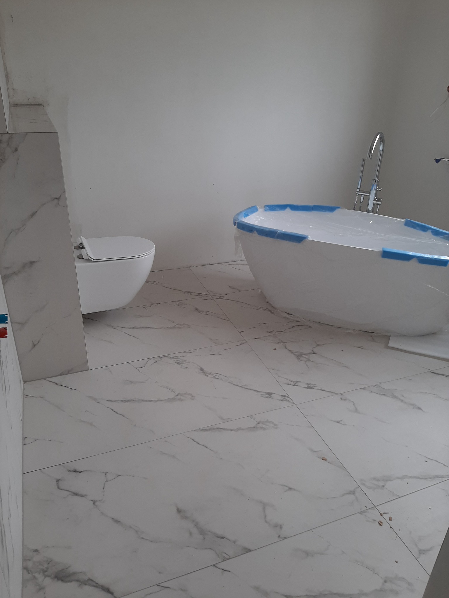 Nowa, wolnostojąca wanna owalna zabezpieczona folią, wisząca toaleta i płytki imitujące marmur w trakcie remontu łazienki.