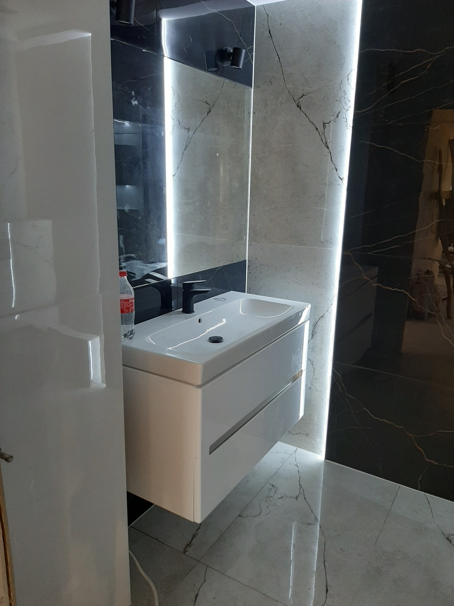 Nowoczesna łazienka z białym blatem umywalki, czarną armaturą i marmurowymi płytkami z podświetleniem LED. Na blacie umywalki stoi butelka wody.