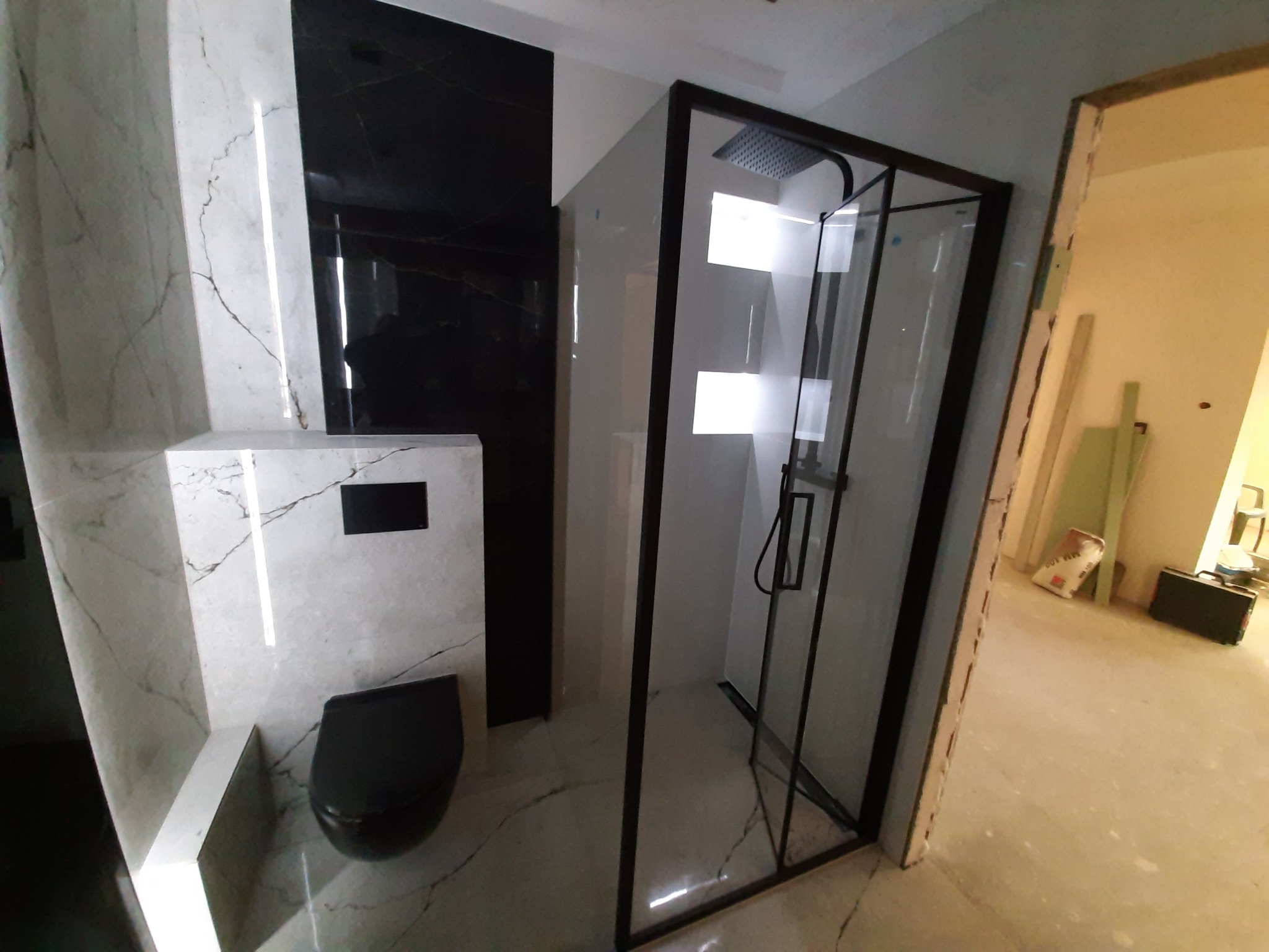 Nowoczesna łazienka w trakcie wykończenia, widoczna czarna armatura, kabina prysznicowa z czarnymi profilami oraz podwieszana toaleta na tle marmurowych płytek.