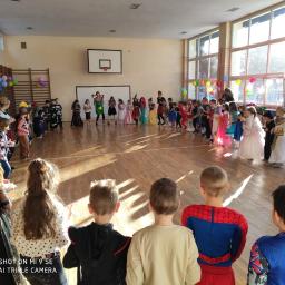 Animacja Sylwia Kojda - Sala gimnastyczna wypełniona dziećmi w kostiumach, ustawionymi w okręgu wokół osoby prowadzącej zabawę; widoczne kolorowe balony i naturalne światło wpadające przez okna.