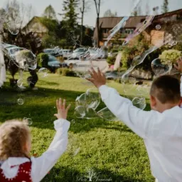 Dwoje dzieci, chłopiec i dziewczynka w ludowym stroju, bawią się bańkami mydlanymi na zielonym trawniku, w tle budynek.