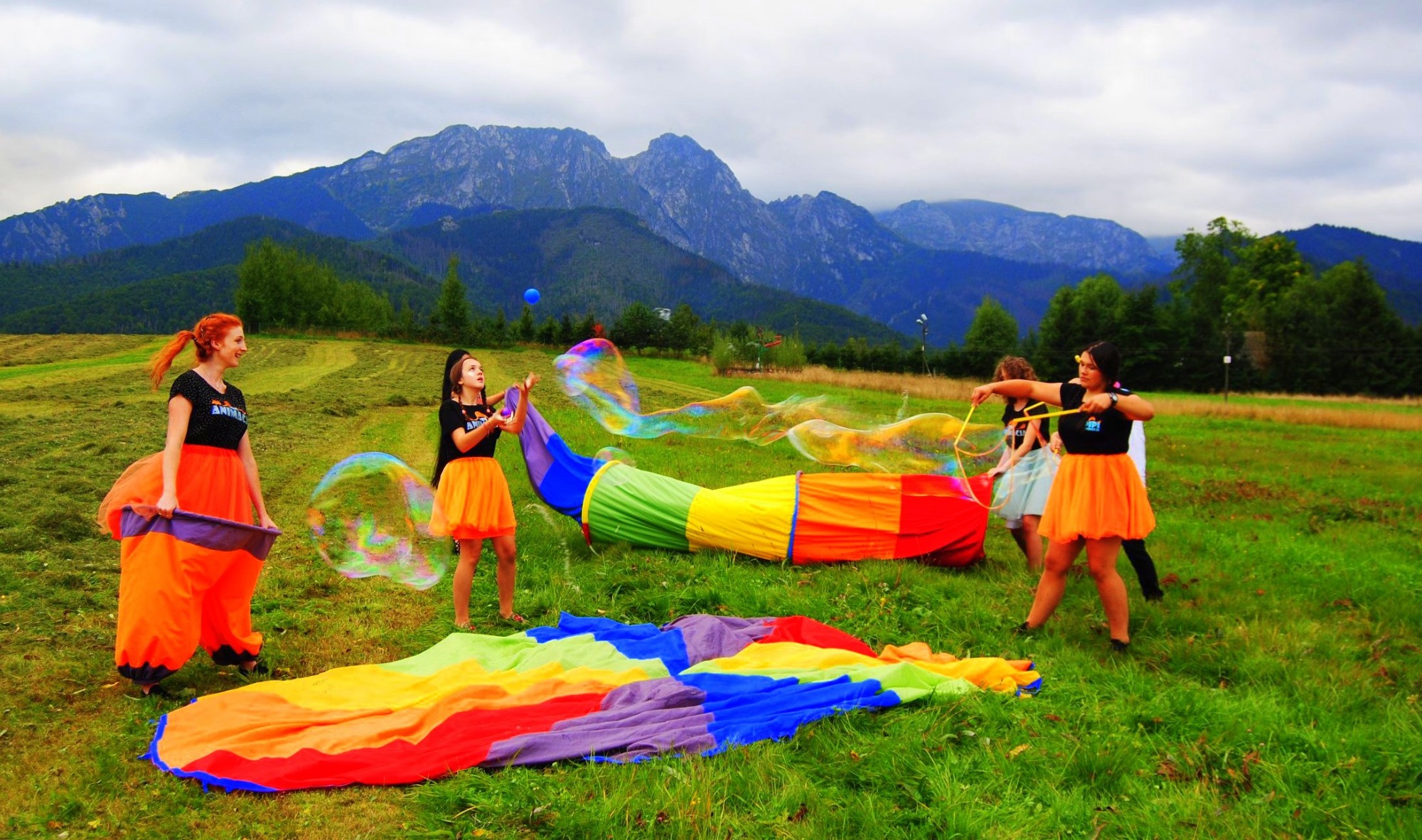 Grupa osób w pomarańczowych spódniczkach puszcza ogromne bańki mydlane na zielonej łące z widokiem na góry w tle. Jedna osoba trzyma kolorowy tunel, a druga używa sznurka do tworzenia baniek.