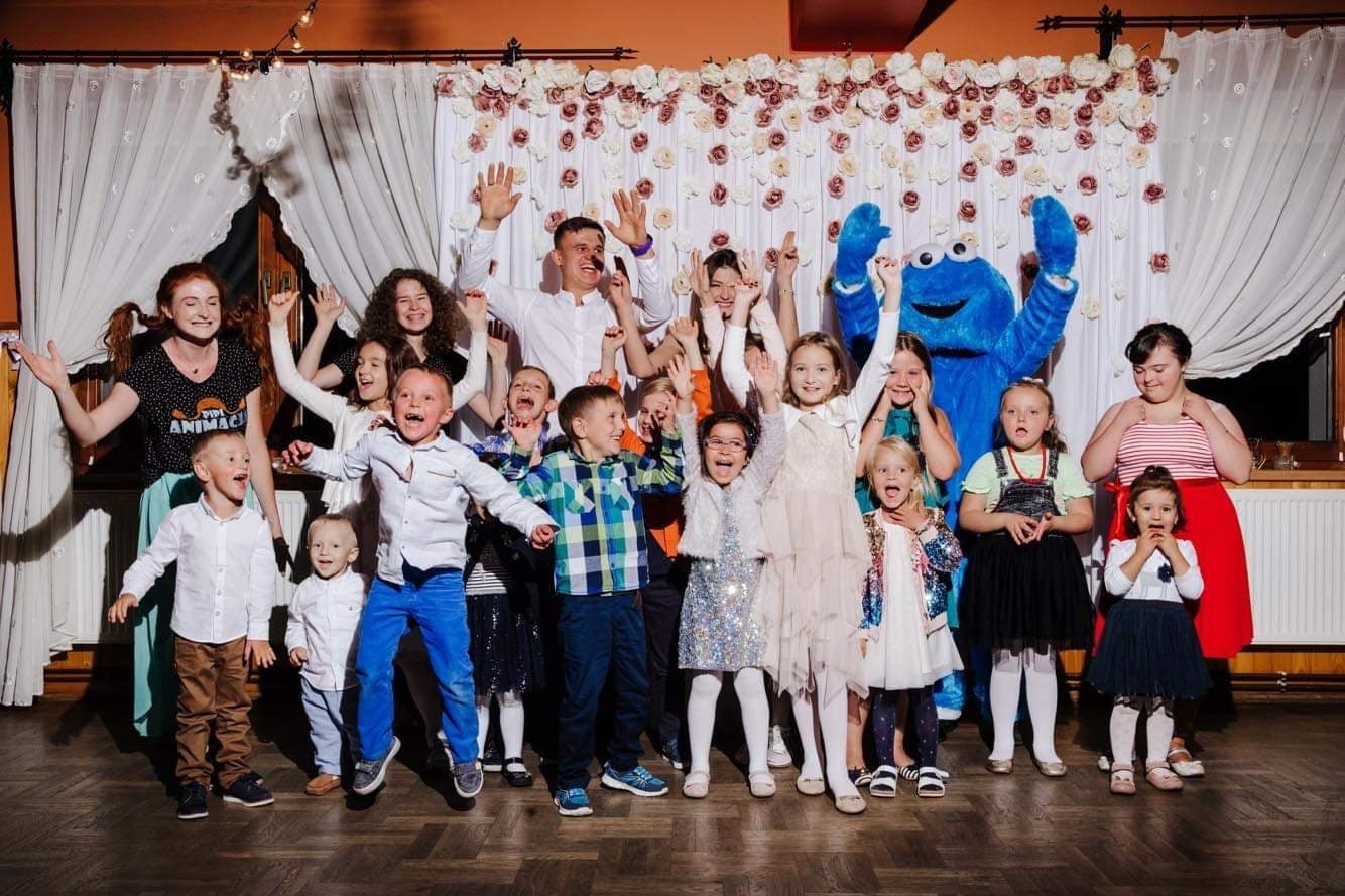 Grupa rozbawionych dzieci i dorosłych, w tym osoba w kostiumie niebieskiego potwora, pozuje na tle dekoracji z białych i różowych kwiatów na imprezie.