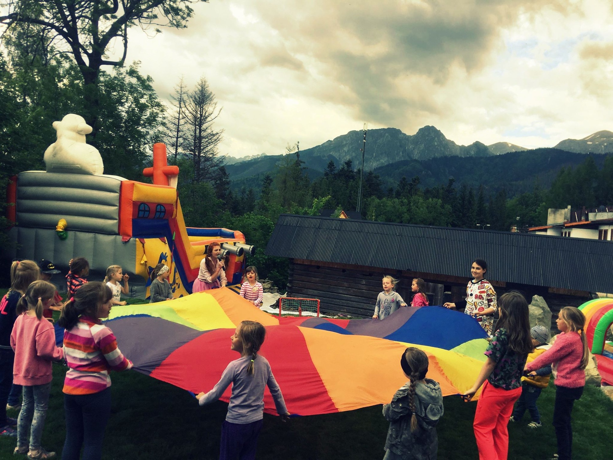 Grupa dzieci bawi się kolorową chustą animacyjną na trawie, w tle dmuchany zamek i góry