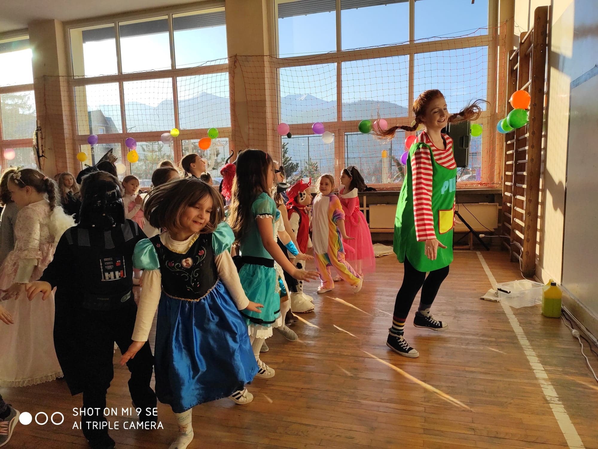 Grupa dzieci w różnorodnych kostiumach, w tym Darth Vader i Elsa, tańczy w sali gimnastycznej, prowadzona przez osobę przebraną za Pippi Pończoszankę, widoczne balony i drabinki gimnastyczne.