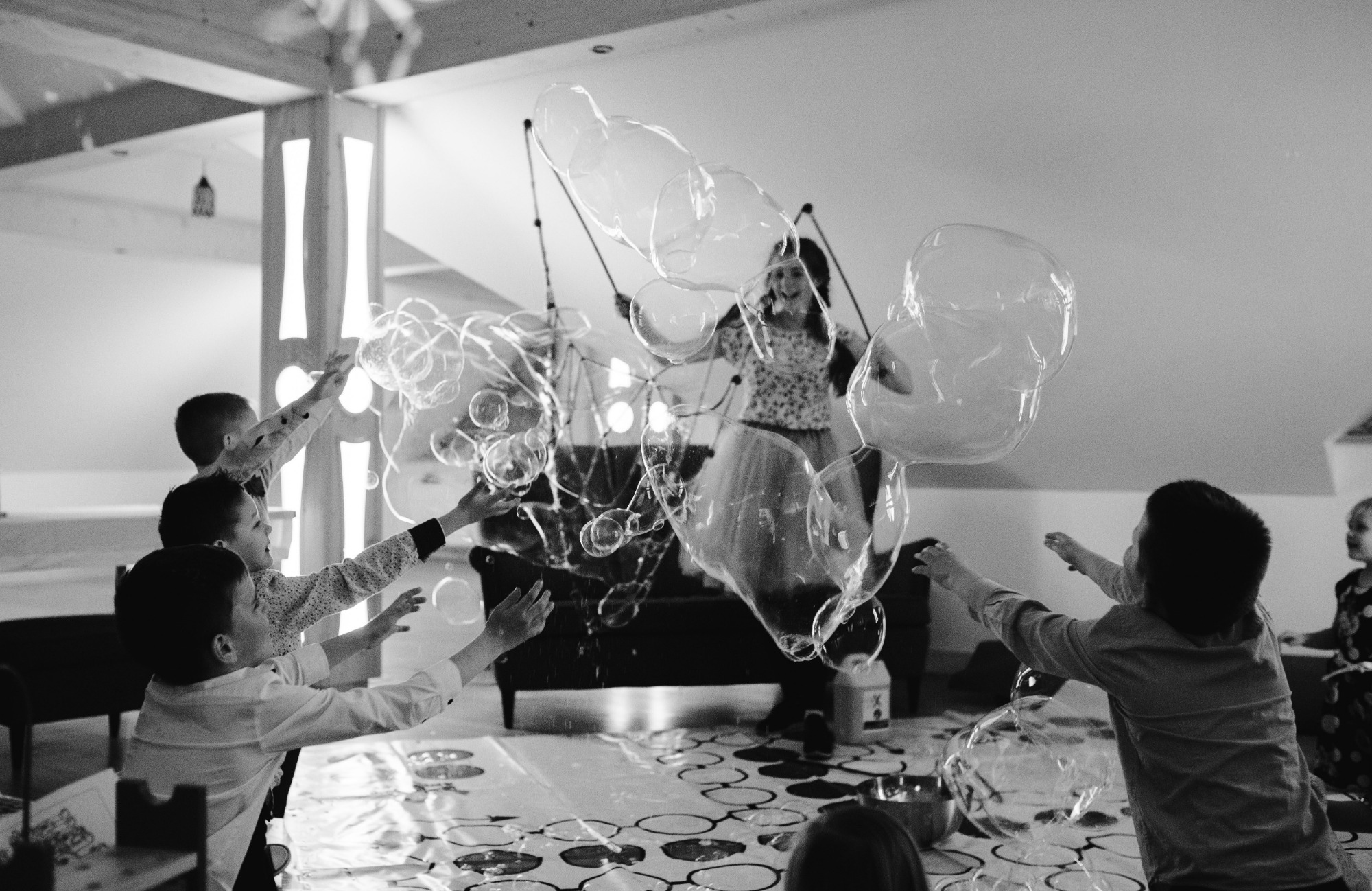 Czarno-białe zdjęcie grupy dzieci próbujących dotknąć ogromnych baniek mydlanych tworzonych przez kobietę z użyciem sznurków i kijków w jasnym pomieszczeniu z drewnianymi elementami konstrukcyjnymi.