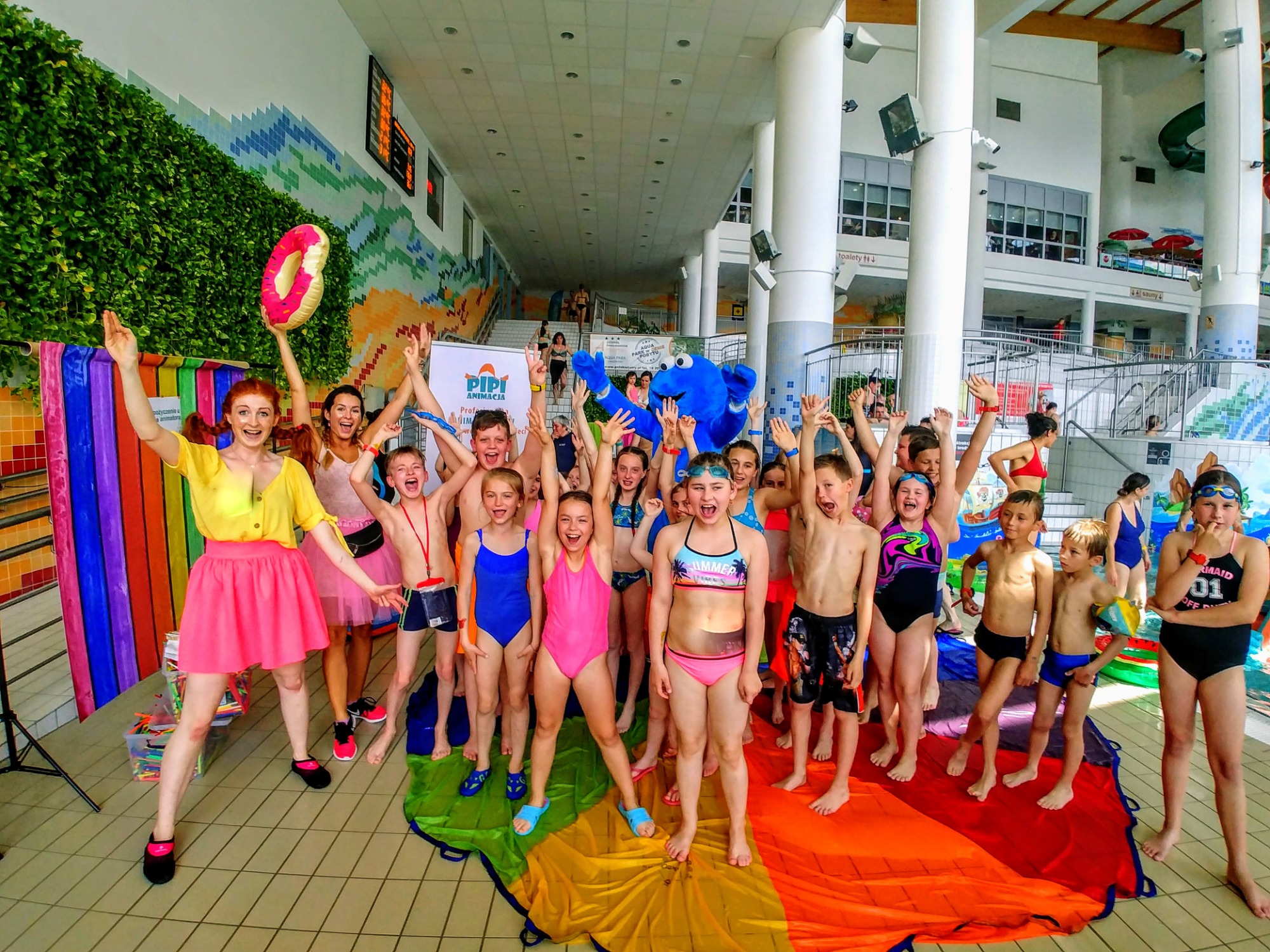 Animator w żółtej bluzce i różowej spódnicy prowadzi grupę dzieci w strojach kąpielowych na tle basenu, z dmuchanym pączkiem w ręku i kolorową płachtą na podłodze, obok stoi osoba w kostiumie...