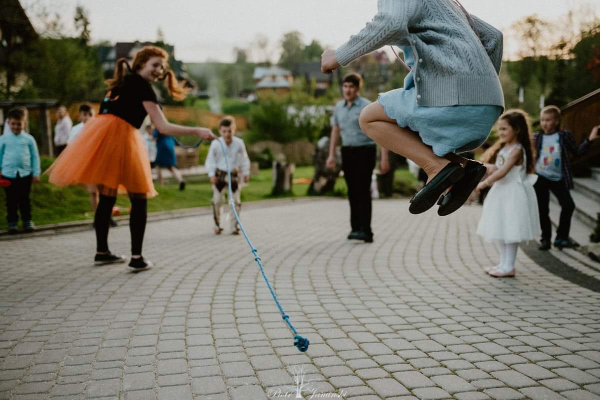 Dziewczynka w locie przeskakuje przez sznur, trzymany przez animatorkę w pomarańczowej spódnicy tiulowej, na tle grupy dzieci i budynków.
