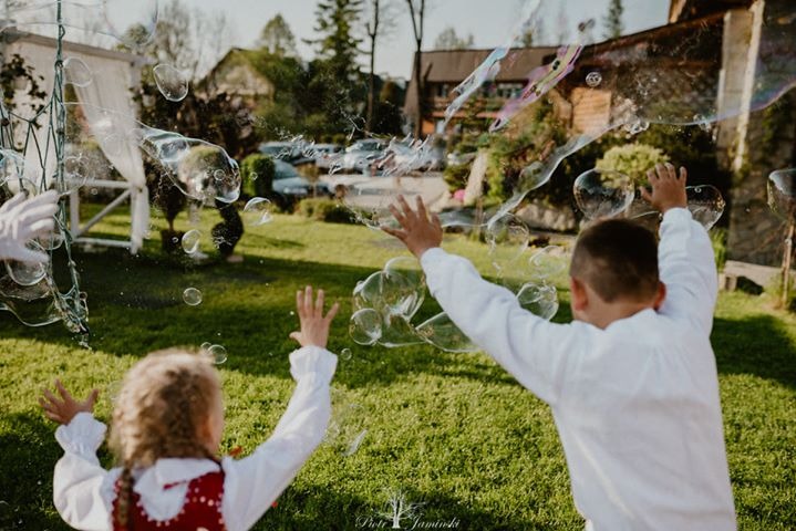 Dwoje dzieci, chłopiec i dziewczynka w ludowym stroju, bawią się bańkami mydlanymi na zielonym trawniku, w tle budynek.