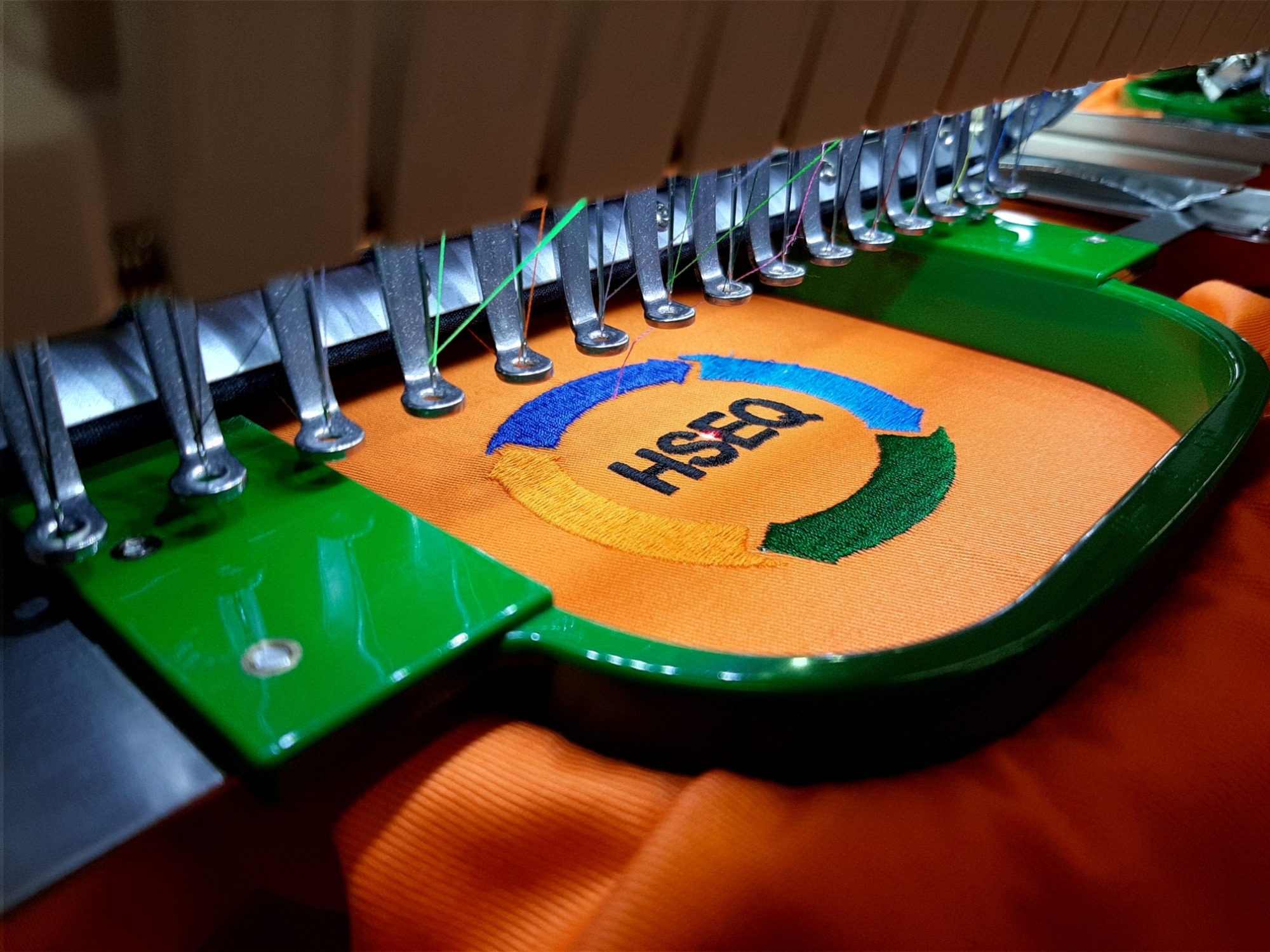 Proces haftowania logo 'HSEQ' w kolorach niebieskim, żółtym i zielonym na pomarańczowej tkaninie w Zawadzie, widoczne igły maszyny do haftu i zielona ramka stabilizująca materiał.
