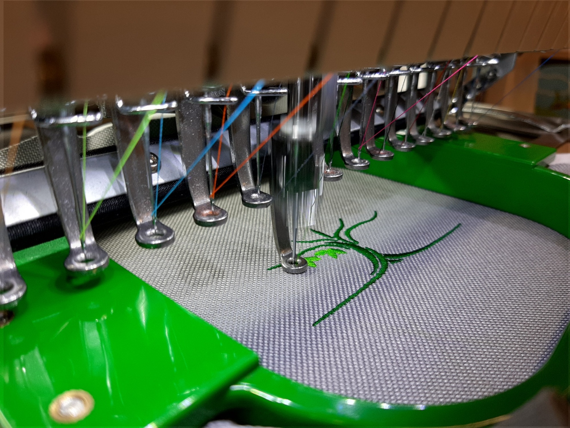 Proces haftowania zielonego wzoru na szarej tkaninie za pomocą przemysłowej maszyny do haftu, widoczne igły i kolorowe nici.